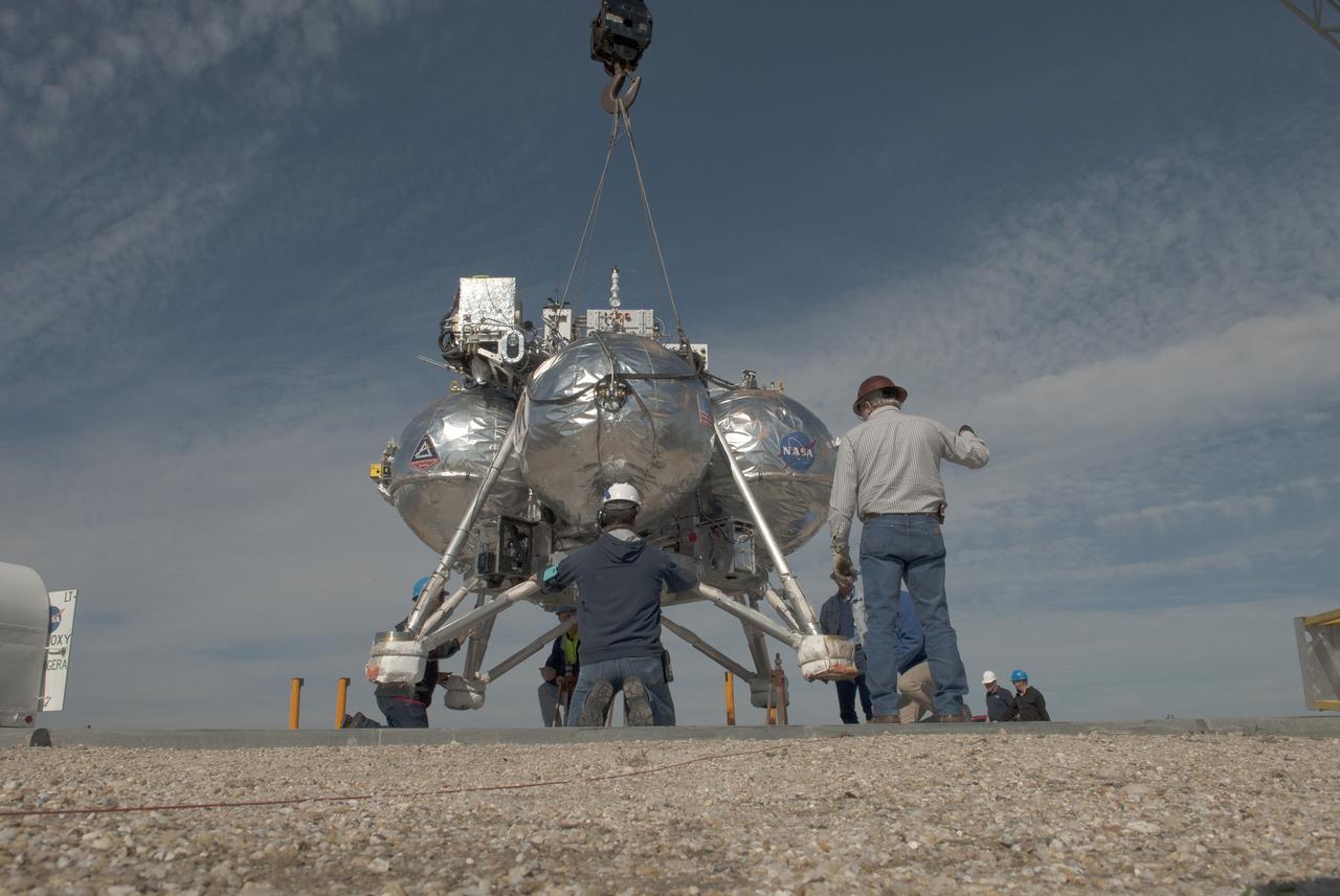 CAPE CANAVERAL, Fla. – Engineers and technicians prepare NASA's Project Morpheus prototype lander for free flight test number 15 on a launch pad at the north end of the Shuttle Landing Facility at NASA's Kennedy Space Center in Florida. Morpheus is being lowered by crane onto the launch pad. The lander will take off from the ground over a flame trench and use its autonomous landing and hazard avoidance technology, or ALHAT sensors, to survey the hazard field to determine safe landing sites. Project Morpheus tests NASA’s ALHAT and an engine that runs on liquid oxygen and methane, which are green propellants. These new capabilities could be used in future efforts to deliver cargo to planetary surfaces. Project Morpheus is being managed under the Advanced Exploration Systems, or AES, Division in NASA’s Human Exploration and Operations Mission Directorate. For more information on Project Morpheus, visit http://morpheuslander.jsc.nasa.gov/.  Photo credit: NASA/Jim Grossmann
