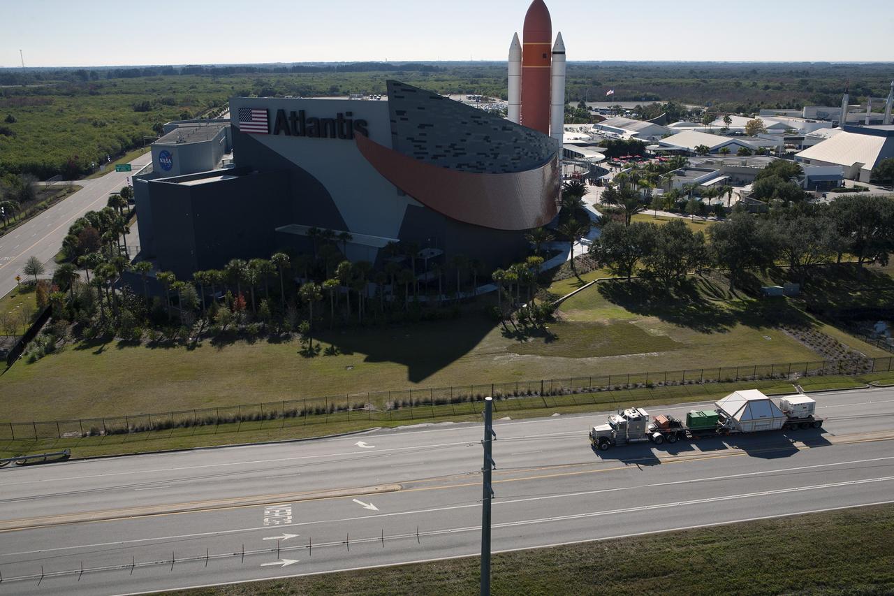 CAPE CANAVERAL, Fla. -- In this aerial view, NASA's Orion crew module, enclosed in its crew module transportation fixture and secured on a flatbed truck is passing the Kennedy Space Center Visitor Complex on its way to the entrance gate to Kennedy Space Center in Florida. In view is the Space Shuttle Atlantis facility. Orion made the 2,700 mile overland trip from Naval Base San Diego in California. Orion was recovered from the Pacific Ocean after completing a two-orbit, four-and-a-half hour mission Dec. 5 to test systems critical to crew safety, including the launch abort system, the heat shield and the parachute system. The Ground Systems Development and Operations Program led the recovery, offload and transportation efforts. For more information, visit www.nasa.gov/orion. Photo credit: NASA/Kim Shiflett