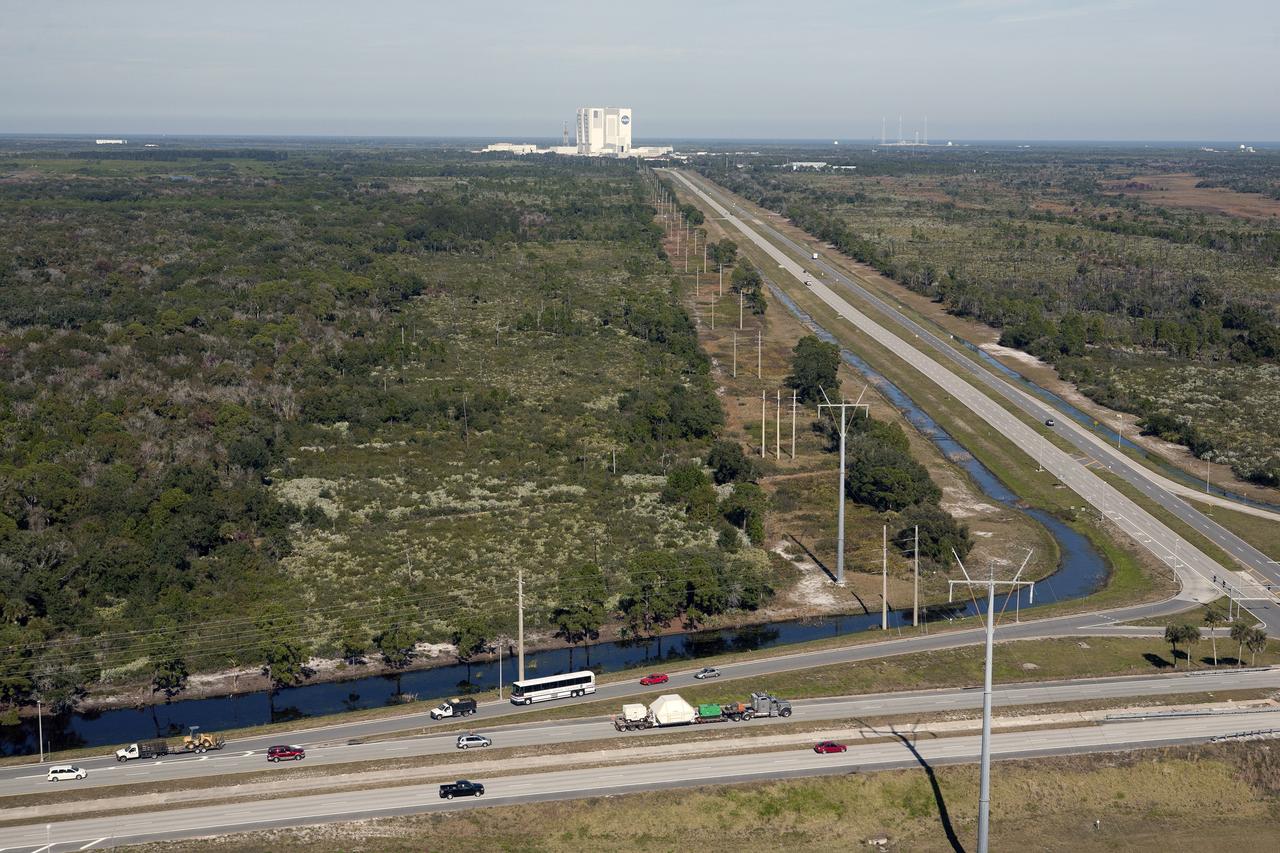 CAPE CANAVERAL, Fla. -- An aerial view reveals the Orion crew module, enclosed in its crew module transportation fixture and secured on a flatbed truck that is proceeding along the NASA Causeway at Kennedy Space Center in Florida. In the background is the iconic Vehicle Assembly Building. Orion made the 2,700 mile overland trip from Naval Base San Diego in California. The spacecraft was recovered from the Pacific Ocean after completing a two-orbit, four-and-a-half hour mission Dec. 5 to test systems critical to crew safety, including the launch abort system, the heat shield and the parachute system. The Ground Systems Development and Operations Program led the recovery, offload and transportation efforts. For more information, visit www.nasa.gov/orion. Photo credit: NASA/Kim Shiflett