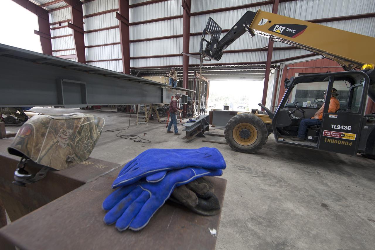 Bright-colored work gloves in the foreground are a stark contrast to the construction work going on at Sauer in Oak Hill, Florida. A forklift is being used to lift a segment of the first of 10 new work platforms for high bay 3 in the Vehicle Assembly Building at NASA's Kennedy Space Center in Florida, for buildup. A contract to modify high bay 3 was awarded by NASA to the Hensel Phelps Construction Co. of Orlando, Florida in March 2014. Sauer is a subcontractor to Hensel Phelps. The Ground Systems Development and Operations Program is overseeing upgrades and modifications to the high bay to support processing of NASA's Space Launch System and Orion spacecraft, and other exploration vehicles. Photo credit: NASA/Ben Smegelsky