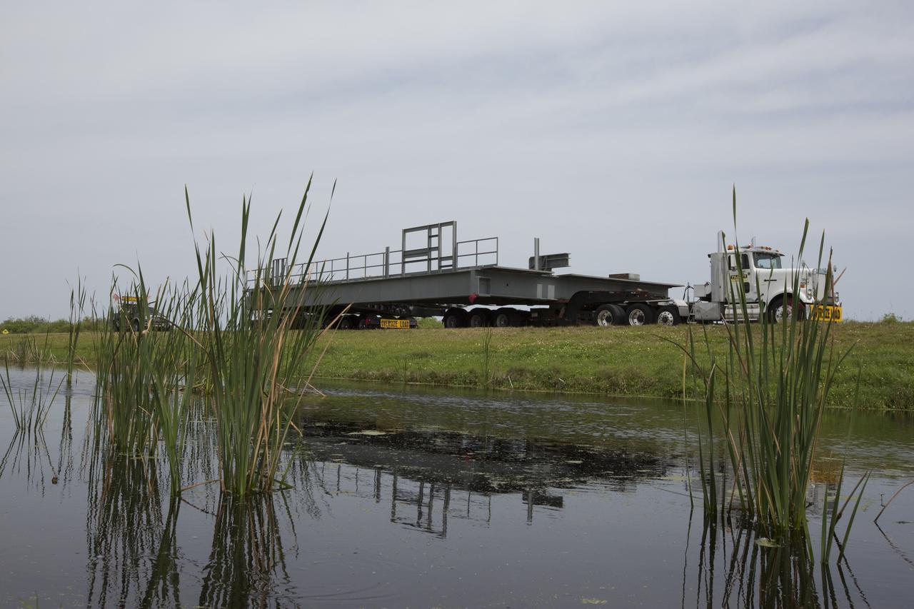 With its image reflected in the water, a heavy load transport truck proceeds along the road to the Vehicle Assembly Building (VAB) at NASA’s Kennedy Space Center in Florida, carrying the second half of the F-level work platforms for the agency’s Space Launch System (SLS) rocket. The platform will be delivered to the VAB staging area in the west parking lot. The Ground Systems Development and Operations Program is overseeing upgrades and modifications to VAB High Bay 3 to support processing of the SLS and Orion spacecraft. A total of 10 levels of new platforms, 20 platform halves altogether, will surround the SLS rocket and Orion spacecraft and provide access for testing and processing. Delivery of this platform brings the total to 10 platforms, or half of the work platforms delivered to Kennedy, 