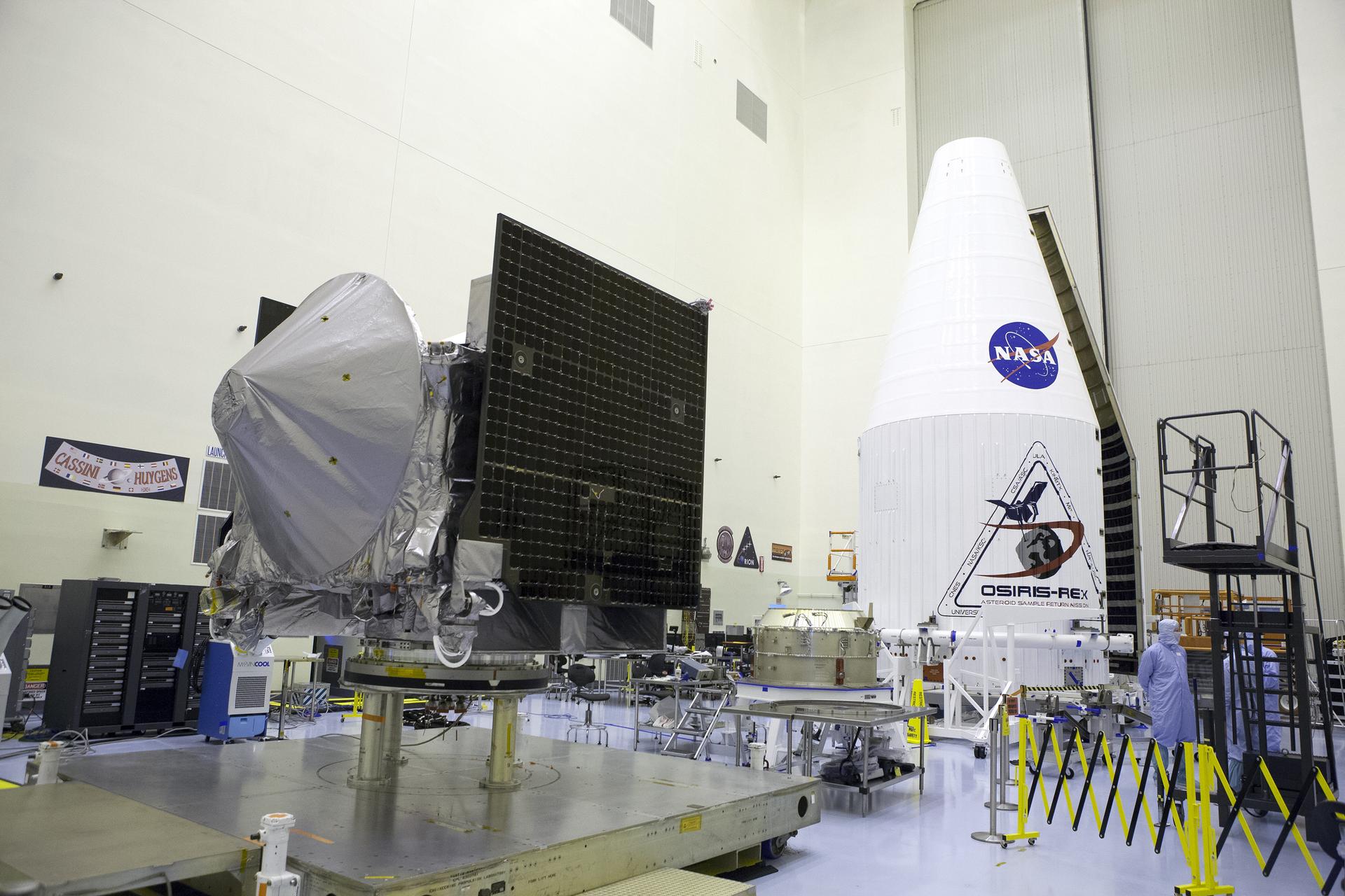The OSIRIS-REx spacecraft sits on a platform inside a clean room. It is a silver rectangle, with a squat silver cone on the front, like a shield. Square black solar panels are attached on either side, folded up like wings. The rocket cone that will hold the spacecraft sits in the background.
