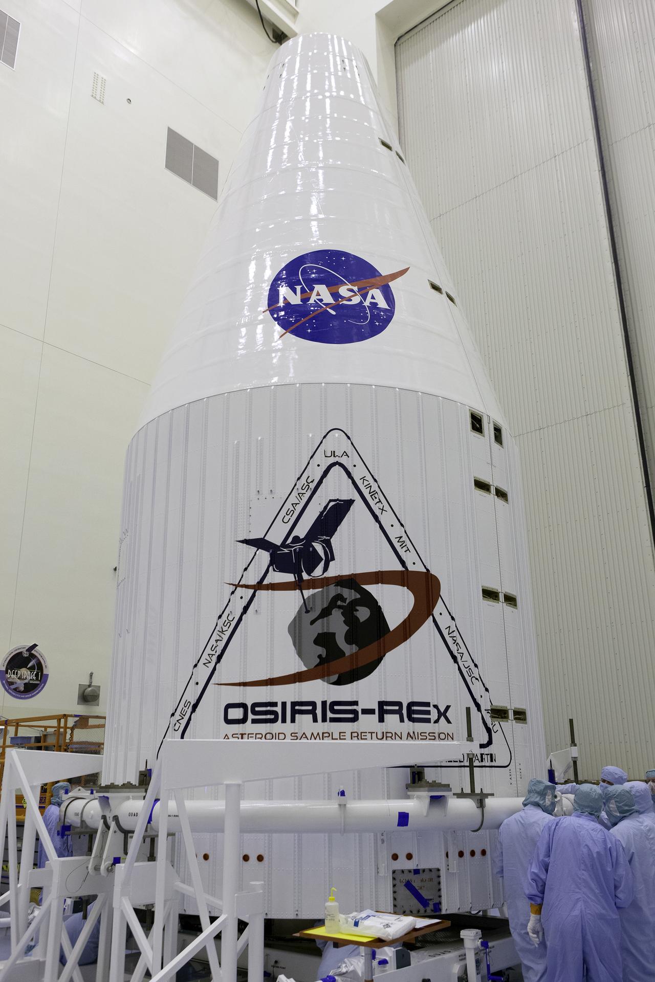 The white rocket nose cone holding the OSIRIS-REx spacecraft. The blue and red NASA meatball logo is painted on the pointed part of the cone. The OSIRIS-REx mission logo is painted on the cylindrical part.