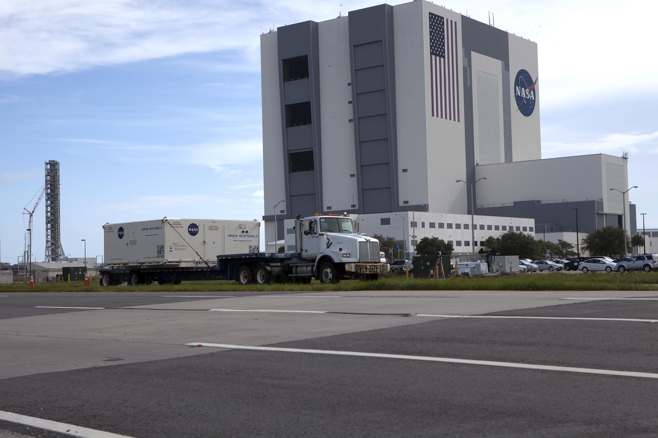 A flatbed truck with the shipping container carrying the Orion heat shield for Exploration Mission 1 (EM-1) passes by the Vehicle Assembly at NASA’s Kennedy Space Center in Florida. The heat shield arrived aboard the agency’s Super Guppy aircraft at the Shuttle Landing Facility, managed and operated by Space Florida, from Lockheed Martin’s manufacturing facility near Denver. The heat shield is being transported to the Neil Armstrong Operations and Checkout Building high bay for processing. The Orion spacecraft will launch atop NASA’s Space Launch System rocket on EM-1, an uncrewed test flight, in 2018. 