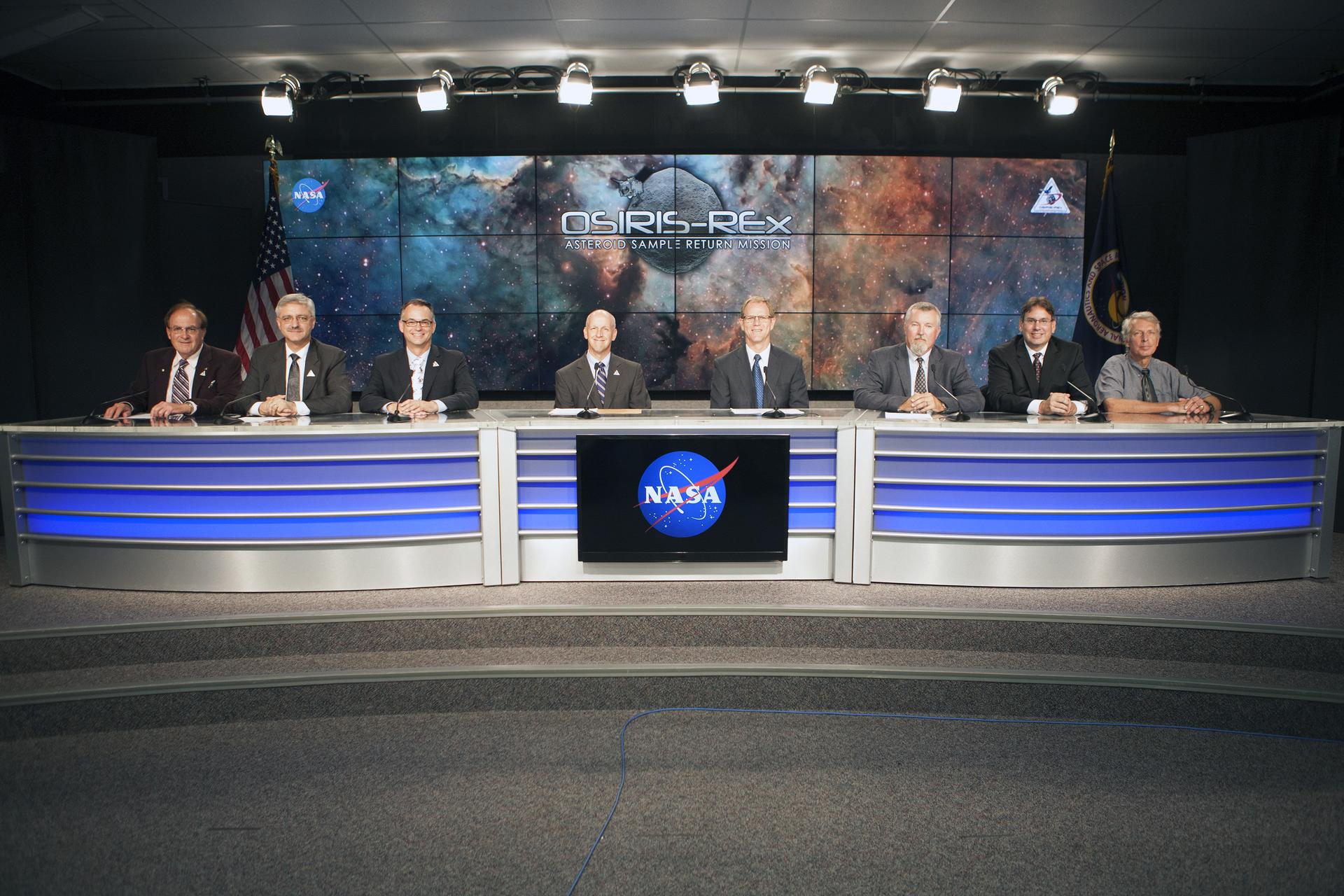 A line of men sit at a long table, in front of an audience. The men are all in suits, with microphones in front of them. A set of screens behind them displays a nebula background with “OSIRIS-REx Asteroid Sample Return Mission” over a generic gray asteroid.