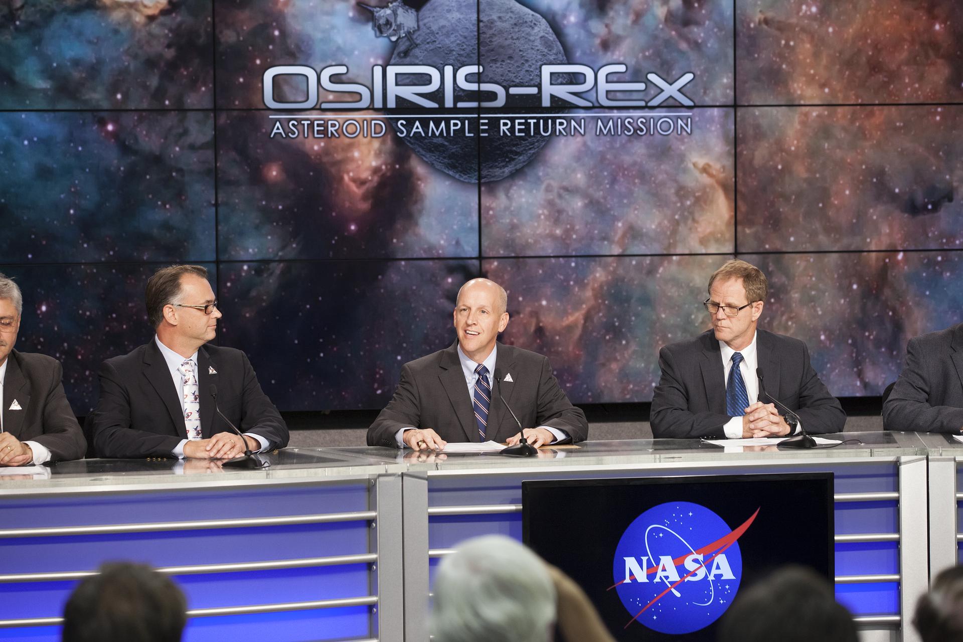 A line of men sit at a long table, silver with blue accents. A screen in the center displays the NASA meatball logo. The men are in suits, with microphones in front of them. The man in the center is in the middle of speaking, and he looks happy. A set of screens behind them displays a nebula background with “OSIRIS-REx Asteroid Sample Return Mission” over a generic gray asteroid.
