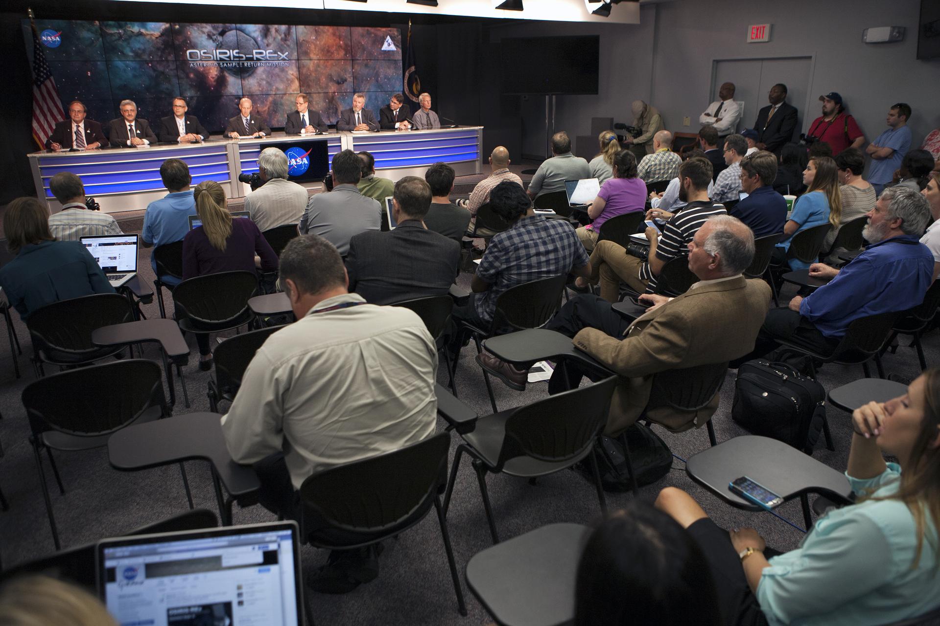 A line of men sit at a long table, in front of an audience. The men are all in suits, with microphones in front of them. The man in the center is in the middle of speaking, and he looks happy. A set of screens behind them displays a nebula background with “OSIRIS-REx Asteroid Sample Return Mission” over a generic gray asteroid.