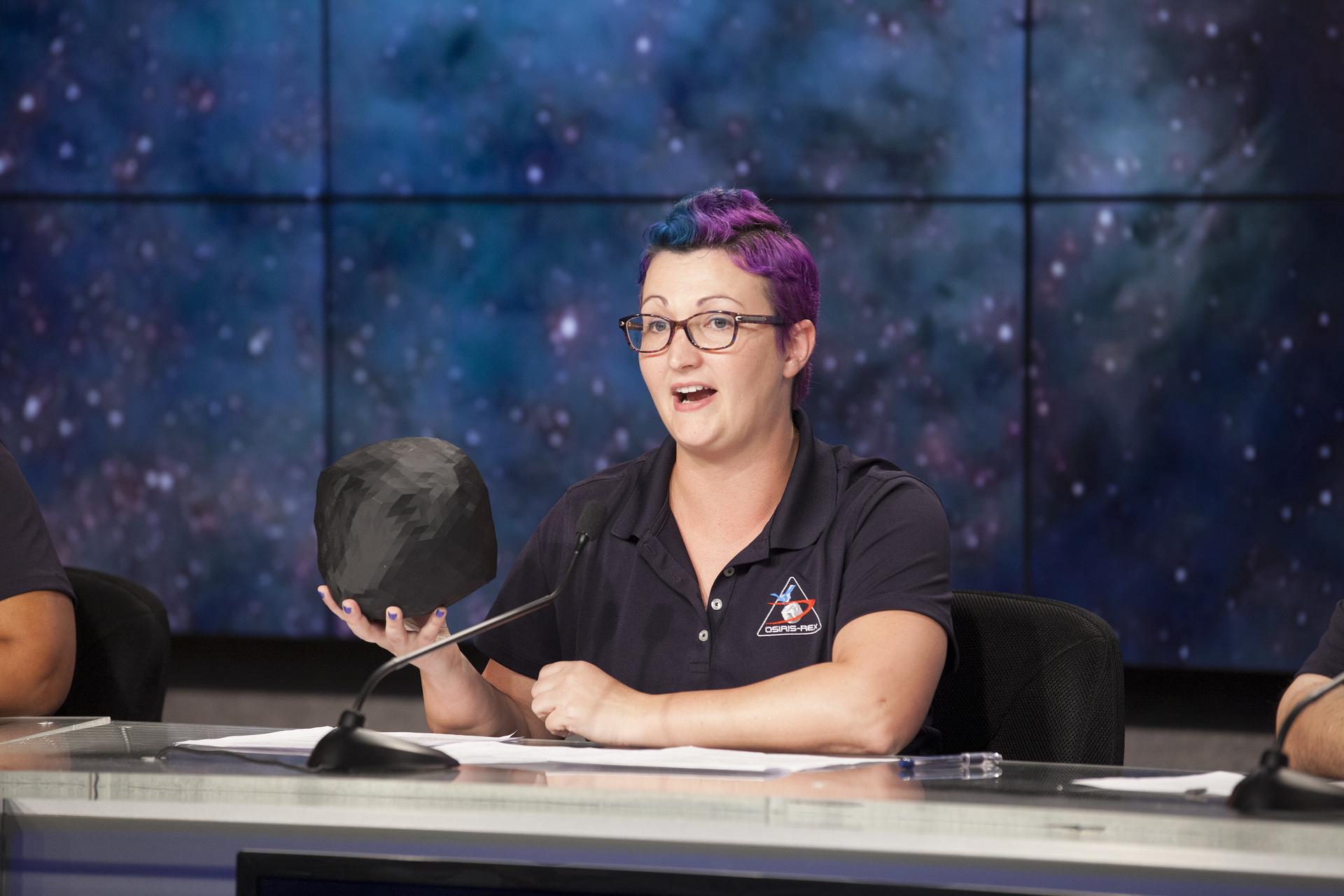 A woman sits behind a table, talking into a microphone. She is holding a 3D printed Bennu and wearing an OSIRIS-REx polo.