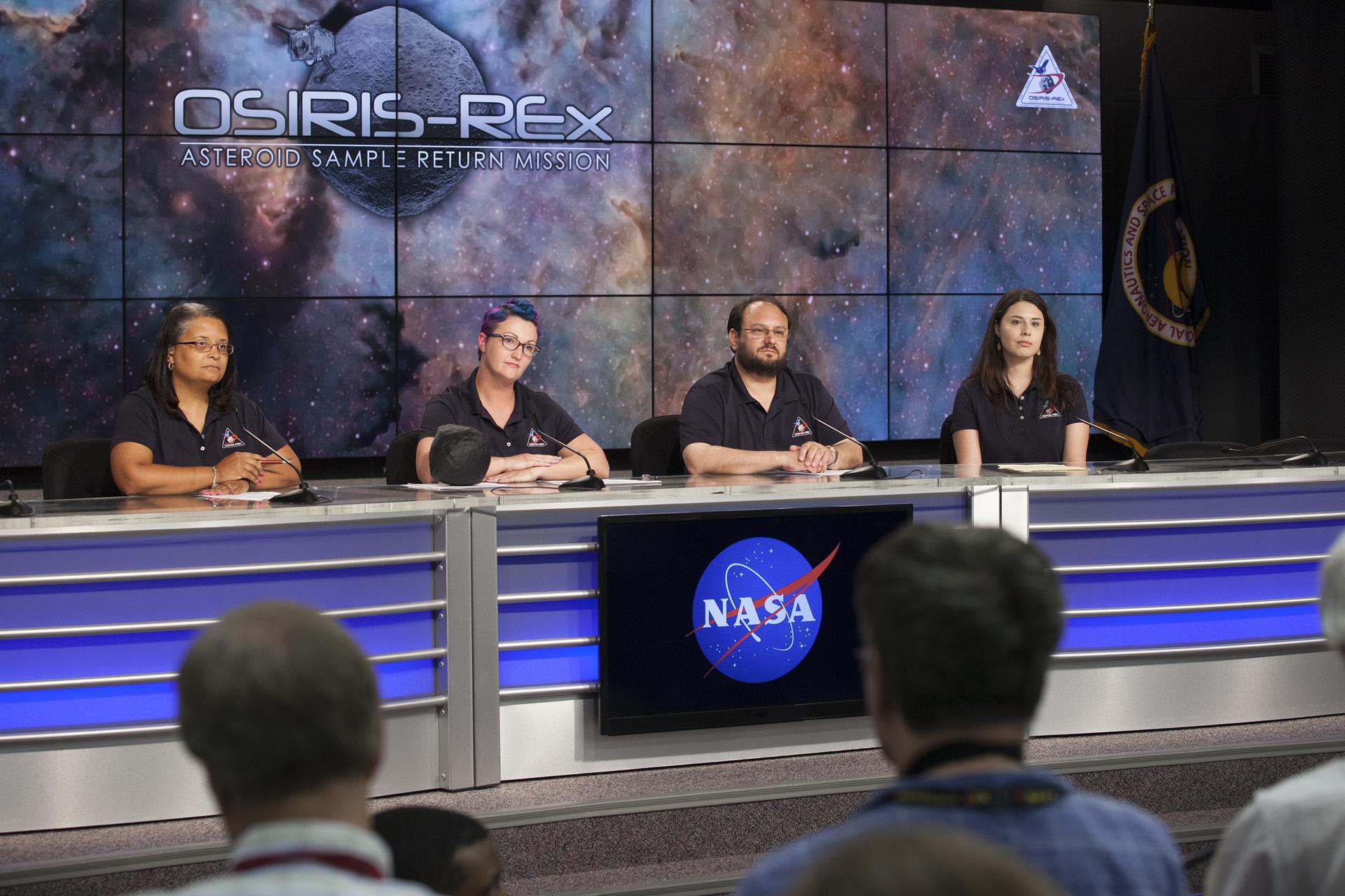 Four people sit in a line at a long table, silver with blue accents. A screen in the center displays the NASA meatball logo. They are all wearing OSIRIS-REx polos, with microphones in front of them. A set of screens behind them displays a nebula background with “OSIRIS-REx Asteroid Sample Return Mission” over a generic gray asteroid.