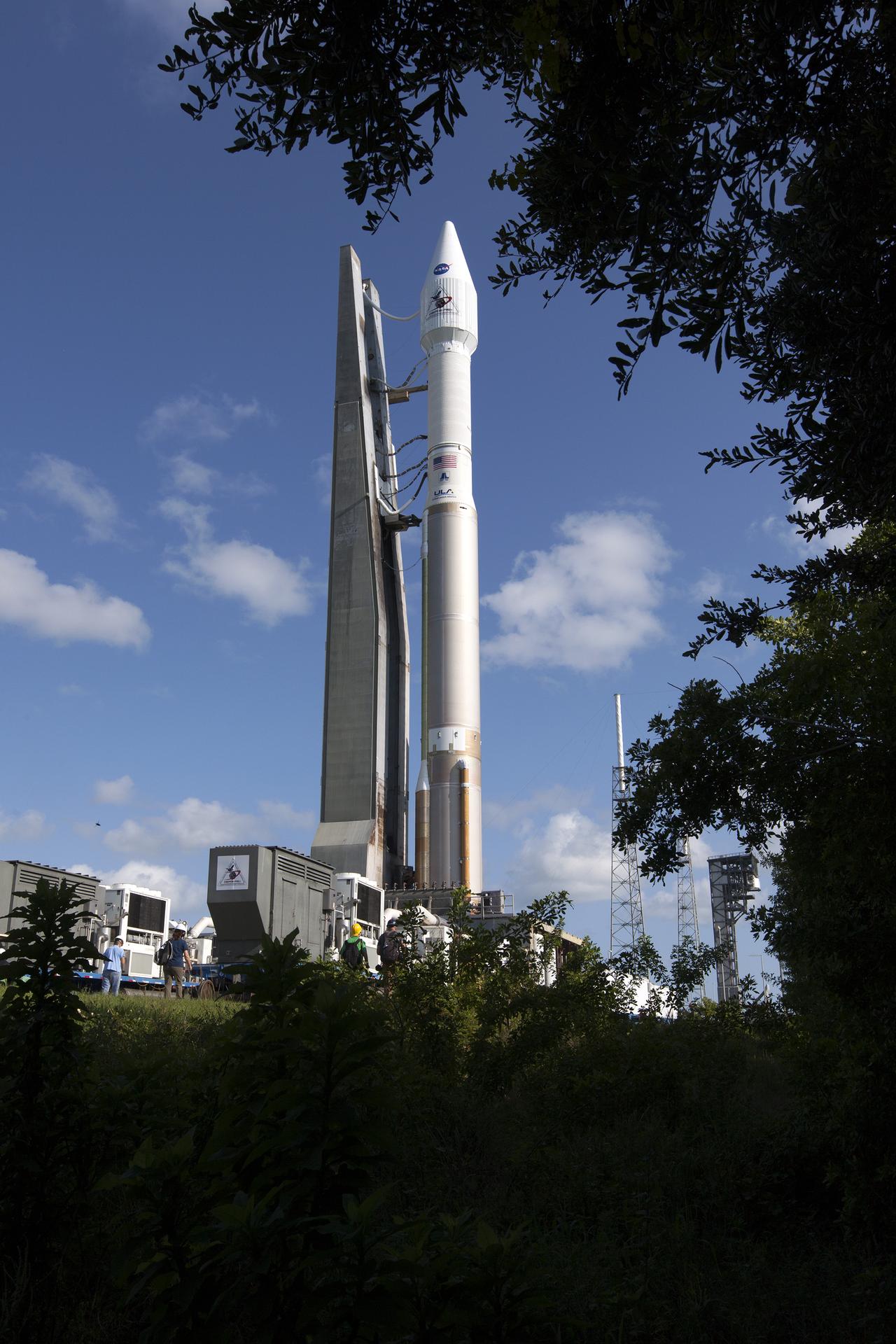 The rocket carrying OSIRIS-REx stands on the launchpad. It is a thin, white rocket against a blue sky with a few clouds.