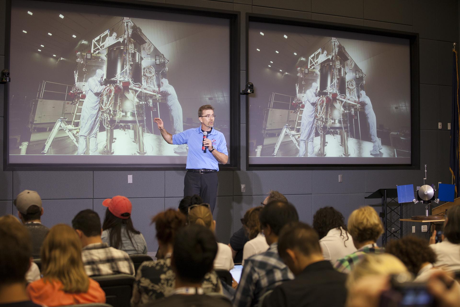 A man talking into a microphone, wearing an OSIRIS-REx polo. A picture of OSIRIS-REx in the clean room is projected on a screen behind him. An audience is visible in front of him.