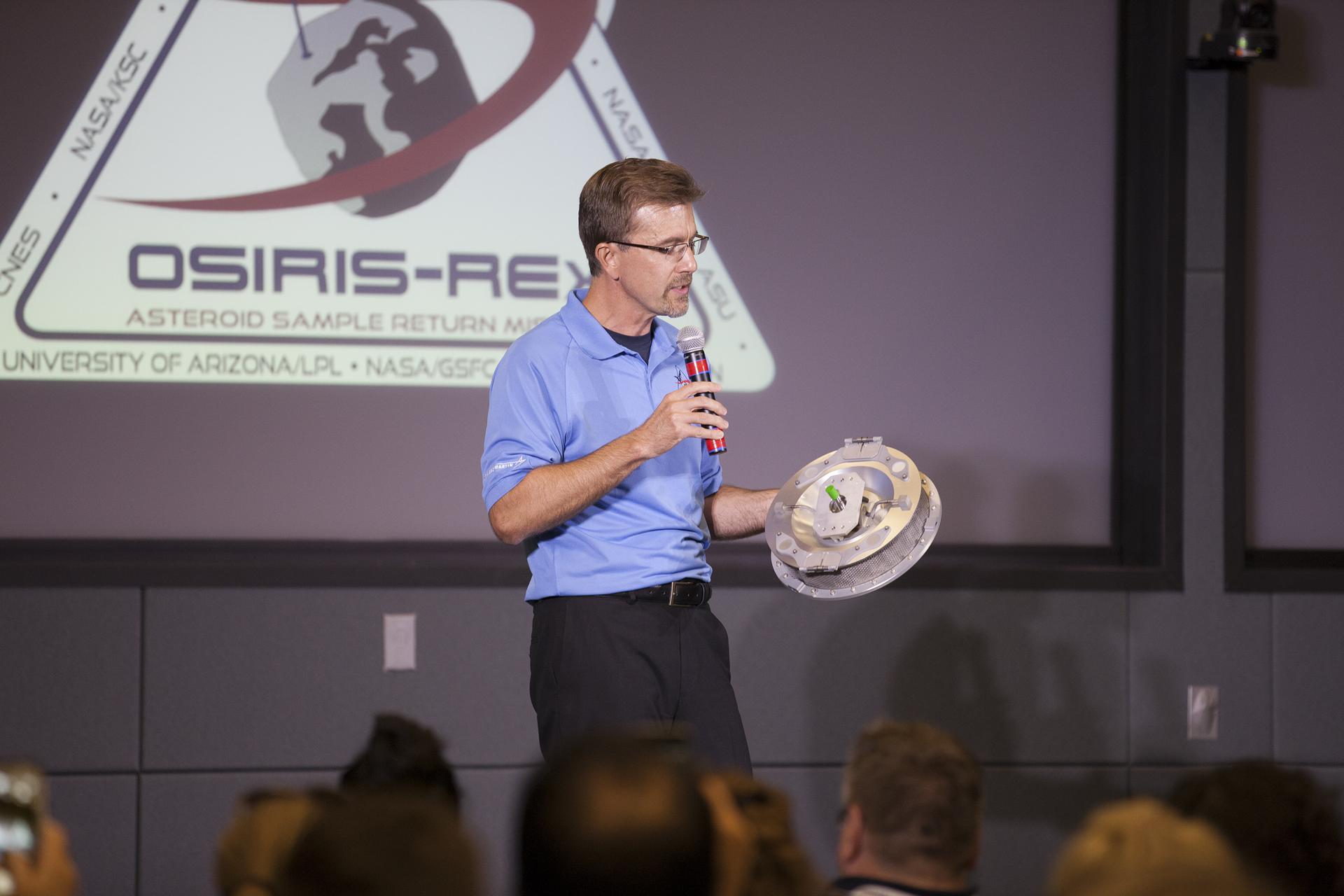 A man talking into a microphone, holding a model of the OSIRIS-REx sample capsule. The OSIRIS-REx logo is projected on a screen behind him. An audience is visible in front of him.