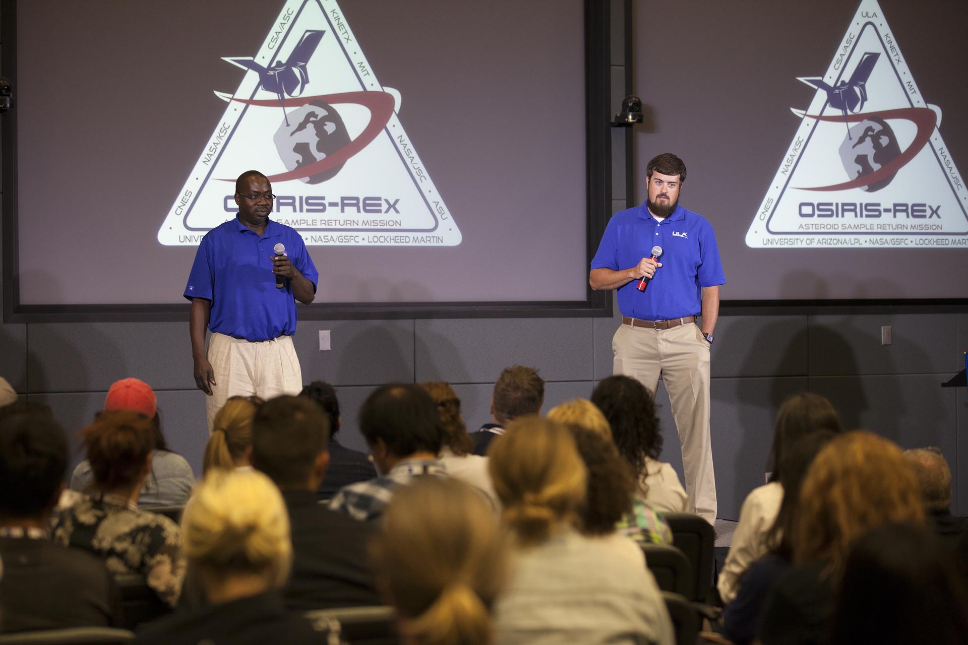 Two men talking into microphones, wearing ULA polos. The OSIRIS-REx logo is projected on multiple screens behind them. An audience is visible in front of them.