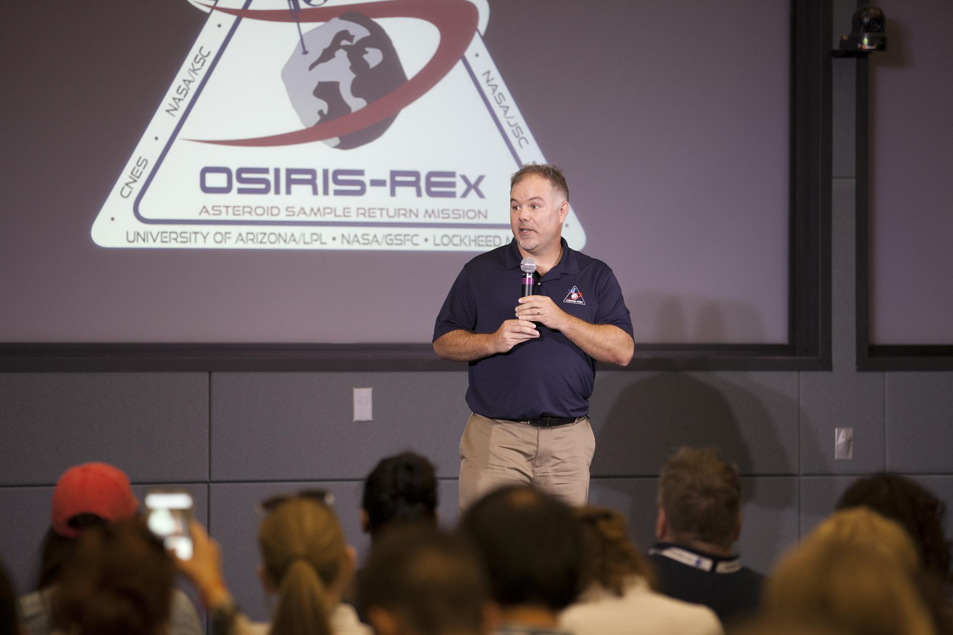 A man talking into a microphone, wearing an OSIRIS-REx polo. The OSIRIS-REx logo is projected on a screen behind him. An audience is visible in front of him.