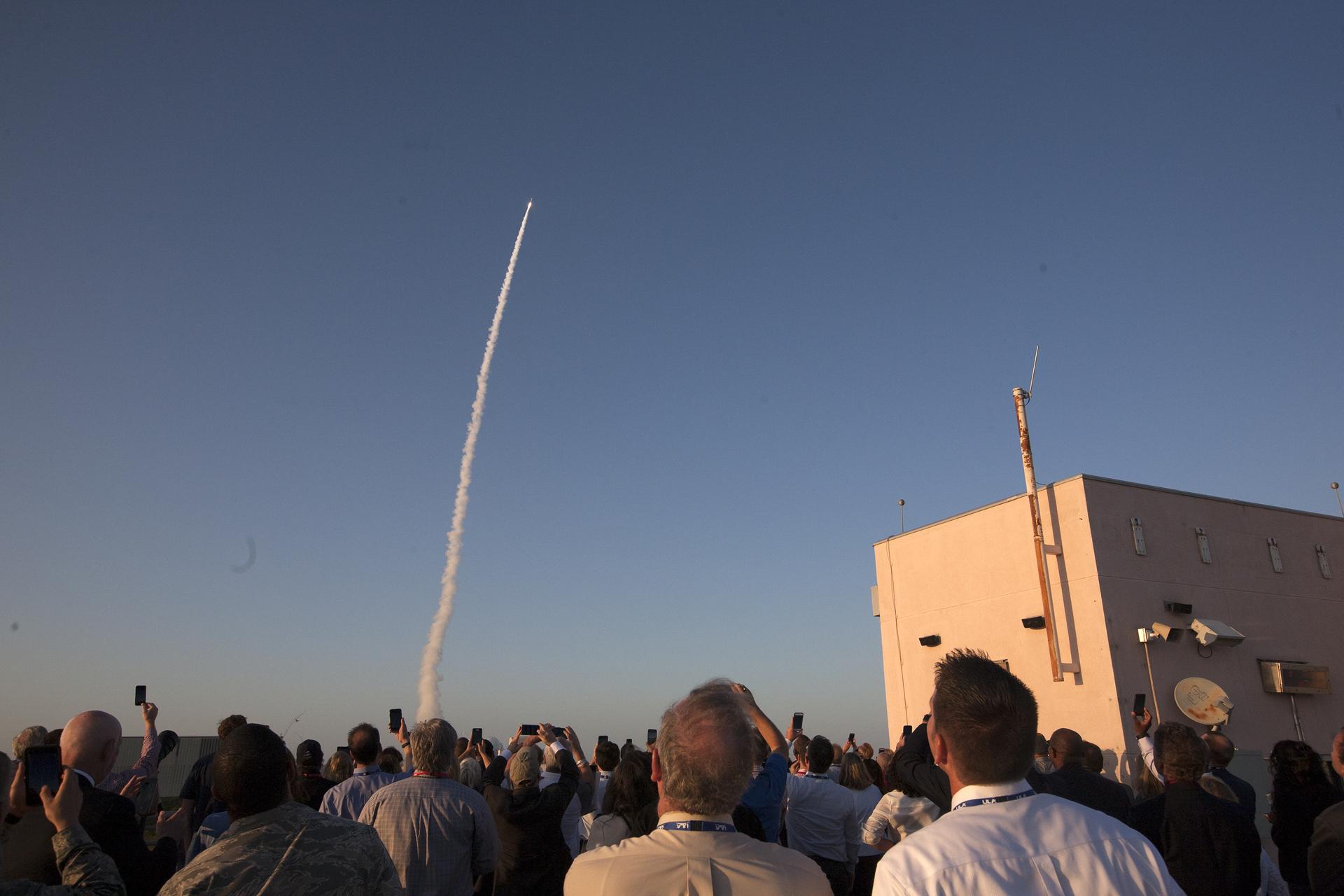 A rocket launching against a light blue sky. It is creating a long trail, curving slightly to the right. A crowd of people are watching.