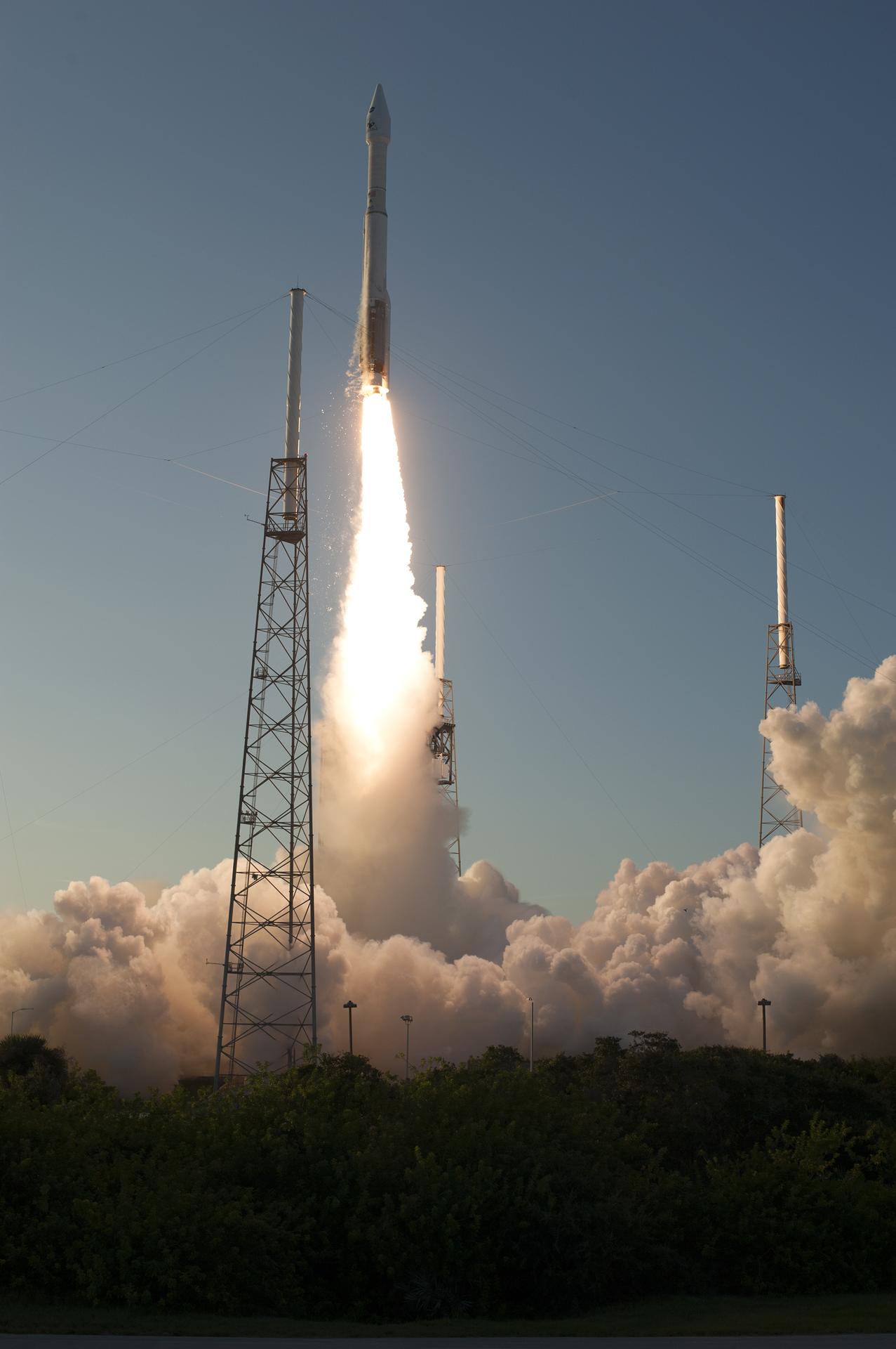 A rocket launching, carrying OSIRIS-REx. The rocket is racing up against a light blue sky, with flames extending out to the ground. A thick cloud of smoke billows across the lower quarter of the image.