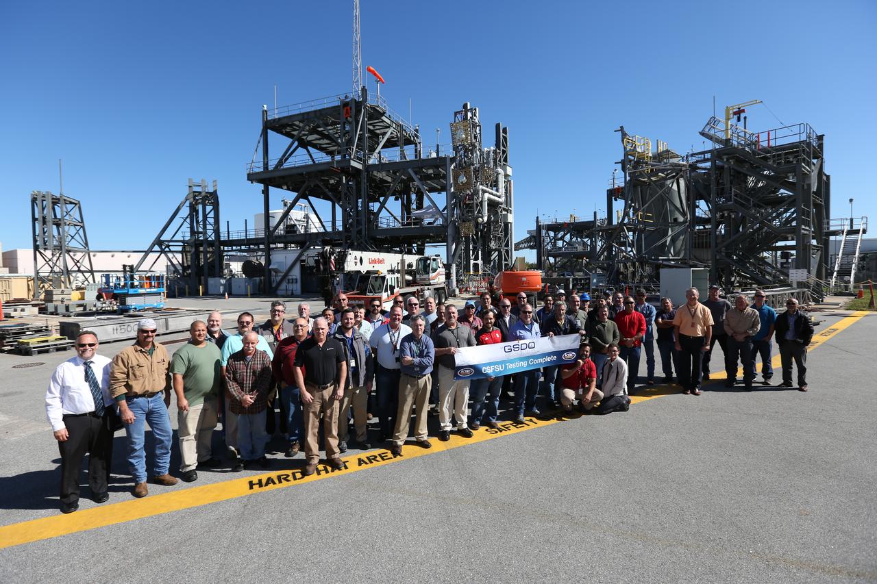 The test team holds a signed banner at the Launch Equipment Test Facility at Kennedy Space Center in Florida. Behind them are some of the test structures used to test the launch umbilicals. Testing of the Core Stage Forward Skirt Umbilical (CSFSU) for NASA's Space Launch System is complete and the umbilical has been transported to the mobile launcher area. The umbilical will be prepared for installation on the tower of the mobile launcher. The CSFSU will be mated to the core stage forward skirt to provide commodities to the SLS rocket, and then disconnect and swing away before launch. Its main purpose is to provide conditioned air and gaseous nitrogen to the SLS Core Stage Forward Skirt. The center’s Engineering Directorate and the Ground Systems Development and Operations Program are overseeing processing and testing of the umbilicals. 