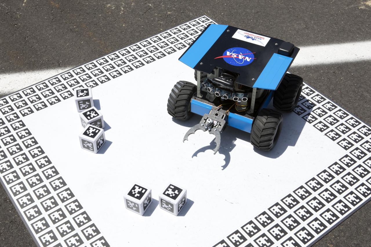 A cube identified with an AprilTag, similar to a barcode, is delivered to a "home" square in the middle of a competition arena during the Swarmathon competition. At the Kennedy Space Center Visitor Complex, student teams developed search algorithms for the Swarmies to operate autonomously, communicating and interacting as a collective swarm similar to ants foraging for food.