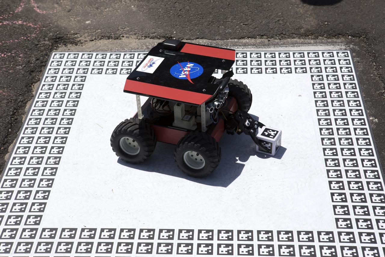 A cube identified with an AprilTag, similar to a barcode, is delivered to a "home" square in the middle of a competition arena during the Swarmathon competition. At the Kennedy Space Center Visitor Complex, student teams developed search algorithms for the Swarmies to operate autonomously, communicating and interacting as a collective swarm similar to ants foraging for food.