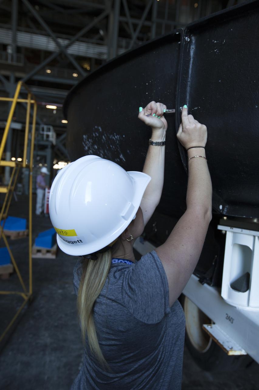 Inside High Bay 2 in the Vehicle Assembly Building (VAB) at NASA's Kennedy Space Center in Florida, a worker helps prepare the Orion heat shield from Exploration Flight Test-1 for unloading off its transporter. The heat shield is being transferred from the Orion Program to the Ground Systems Development and Operations Program, Landing and Recovery Operations. In the VAB, the heat shield will be integrated with the Orion ground test article and used for future underway recovery testing. 