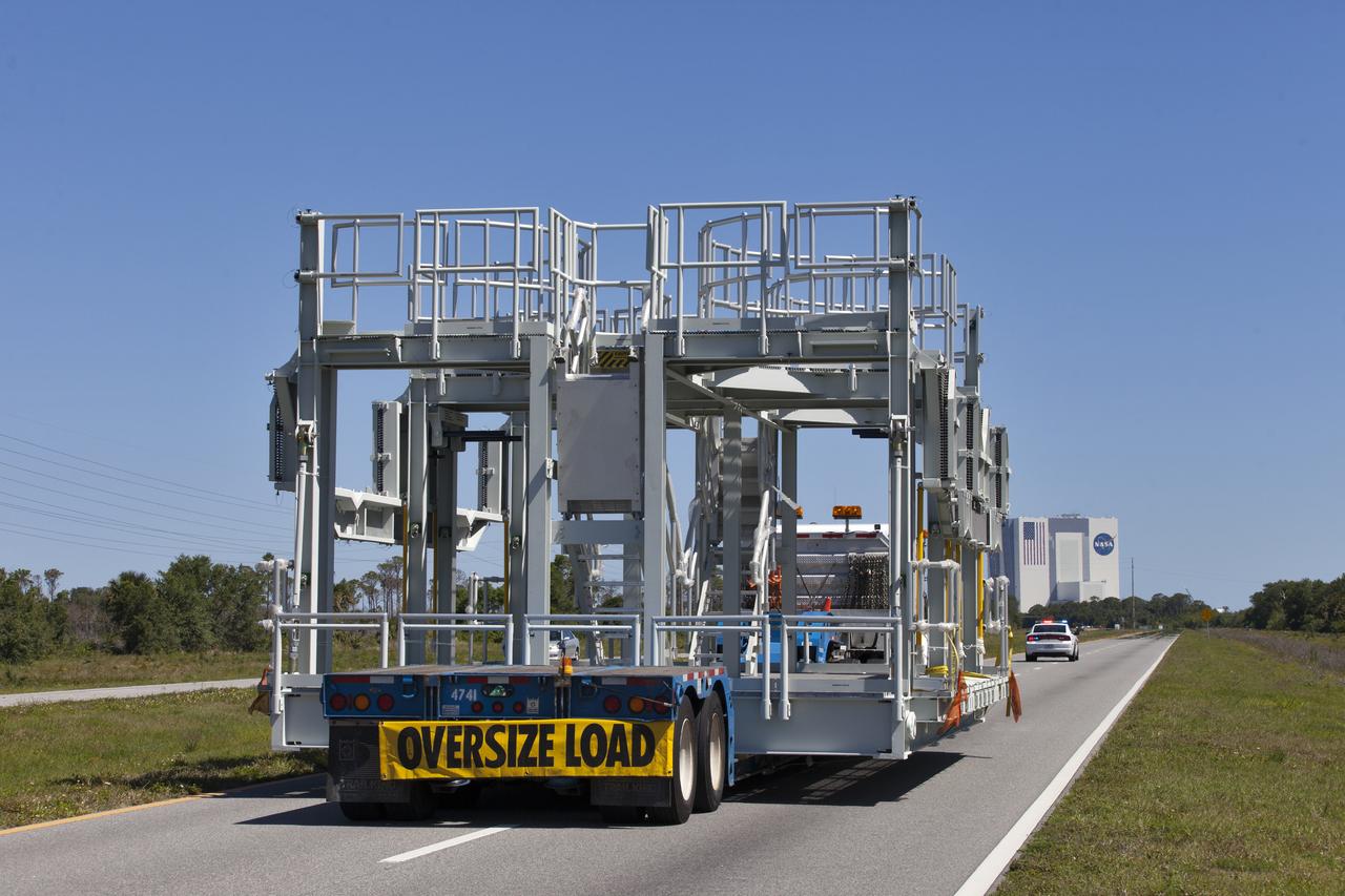 A new service platform for NASA's Space Launch System booster engines, secured on a flatbed truck, is on its way to the Vehicle Assembly Building (VAB), in view in the distance, at the agency's Kennedy Space Center in Florida. It was transported from fabricator Met-Con Inc. in Cocoa, Florida. The platform will be delivered to the VAB, where it will be stored and used for processing and checkout of the engines for the rocket's twin five-segment solid rocket boosters for Exploration Mission-1 (EM-1). During EM-1, an uncrewed Orion spacecraft will launch on the SLS to a stable orbit beyond the Moon and return to Earth for a splashdown in the Pacific Ocean.