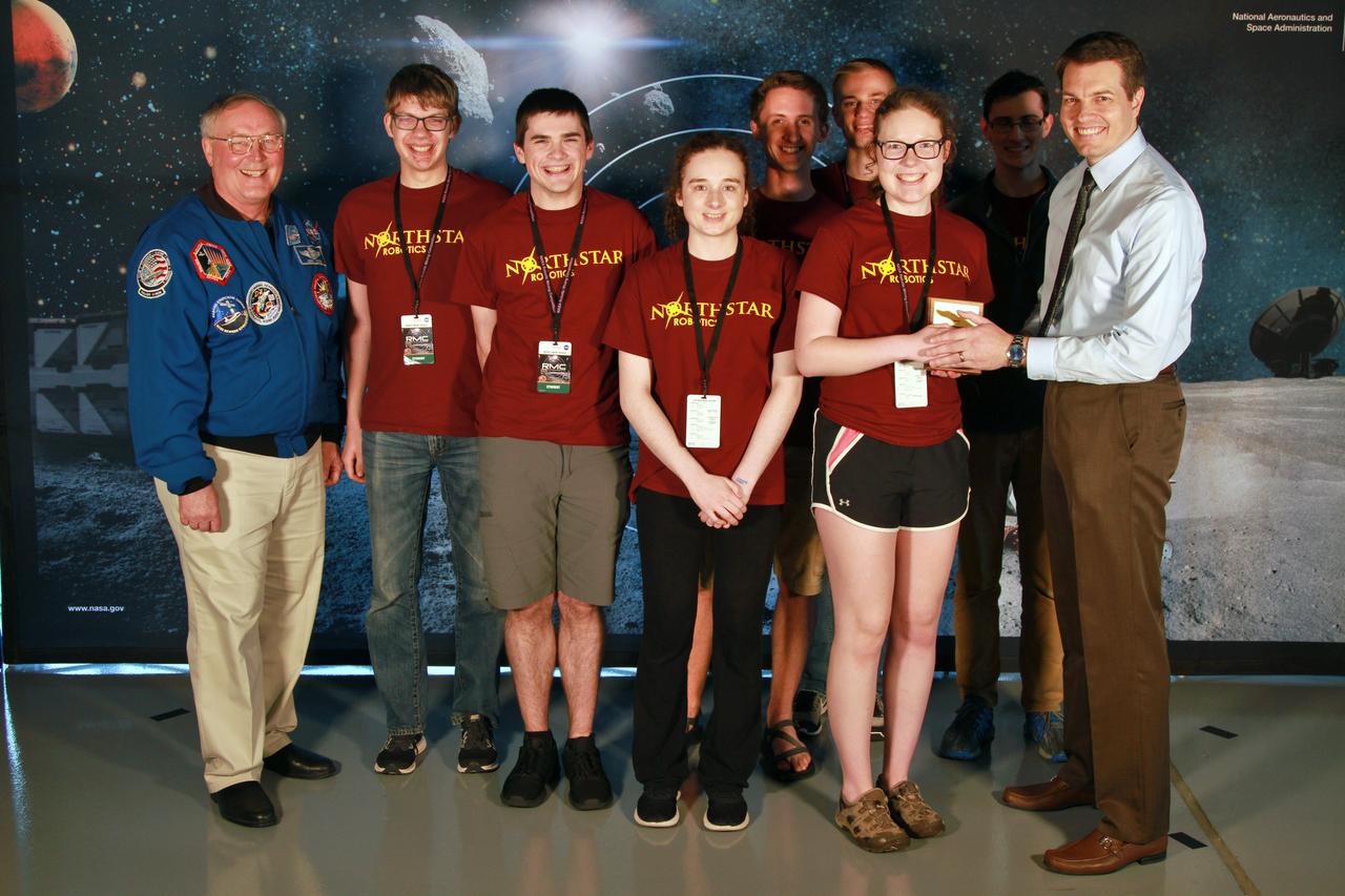 NASA's 9th Annual Robotic Mining Competition concludes with an awards ceremony May 18, 2018, at the Apollo/Saturn V Center at the Kennedy Space Center Visitor Complex in Florida. Team Northstar Robotics from the University of Minnesota - Twin Cities received the Golden E-Stop Award for their robot's safety system. At left is retired NASA astronaut Jerry Ross. At right is Daniel Hull, lead presentation judge. More than 40 student teams from colleges and universities around the U.S. participated in the competition, May 14-18, by using their mining robots to dig in a supersized sandbox filled with BP-1, or simulated lunar soil, gravel and rocks, and participate in other competition requirements. The Robotic Mining Competition is a NASA Human Exploration and Operations Mission Directorate project designed to encourage students in science, technology, engineering and math, or STEM fields. The project provides a competitive environment to foster innovative ideas and solutions that could be used on NASA's deep space missions.