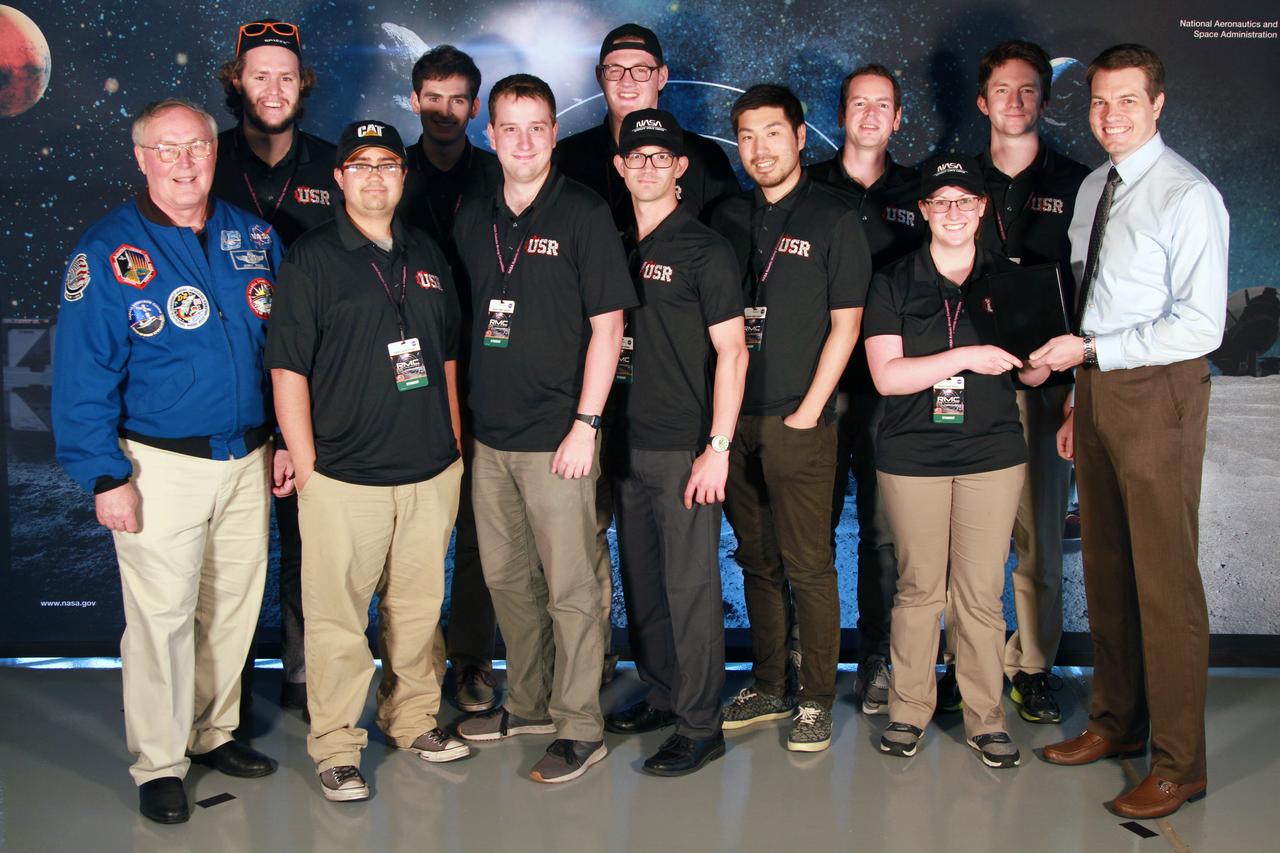 NASA's 9th Annual Robotic Mining Competition concludes with an awards ceremony May 18, 2018, at the Apollo/Saturn V Center at the Kennedy Space Center Visitor Complex in Florida. The Utah Student Robotics team from The University of Utah received second place in the Slide Presentation and Demonstration award category. At left is retired NASA astronaut Jerry Ross. At right is Daniel Hull, lead presentation judge. More than 40 student teams from colleges and universities around the U.S. participated in the competition, May 14-18, by using their mining robots to dig in a supersized sandbox filled with BP-1, or simulated lunar soil, gravel and rocks, and participate in other competition requirements. The Robotic Mining Competition is a NASA Human Exploration and Operations Mission Directorate project designed to encourage students in science, technology, engineering and math, or STEM fields. The project provides a competitive environment to foster innovative ideas and solutions that could be used on NASA's deep space missions.