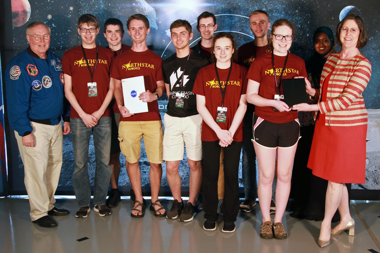 NASA's 9th Annual Robotic Mining Competition concludes with an awards ceremony May 18, 2018, at the Apollo/Saturn V Center at the Kennedy Space Center Visitor Complex in Florida. Team Northstar Robotics from the University of Minnesota - Twin Cities received an honorable mention award for their Systems Engineering Paper. At left is retired NASA astronaut Jerry Ross. At right is Jonette Stecklein, lead systems engineering paper judge. More than 40 student teams from colleges and universities around the U.S. participated in the competition, May 14-18, by using their mining robots to dig in a supersized sandbox filled with BP-1, or simulated lunar soil, gravel and rocks, and participate in other competition requirements. The Robotic Mining Competition is a NASA Human Exploration and Operations Mission Directorate project designed to encourage students in science, technology, engineering and math, or STEM fields. The project provides a competitive environment to foster innovative ideas and solutions that could be used on NASA's deep space missions.