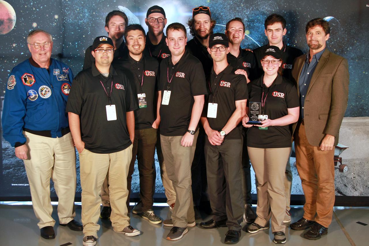 NASA's 9th Annual Robotic Mining Competition concludes with an awards ceremony May 18, 2018, at the Apollo/Saturn V Center at the Kennedy Space Center Visitor Complex in Florida. The Utah Student Robotics team from The University of Utah received The Regolith Mechanics Award for the best example of a real granular innovation that identified a specific regolith mechanics problem and intentionally improved their robot design to deal with it. At left is retired NASA astronaut Jerry Ross. At right is Phil Metzger, mining judge. More than 40 student teams from colleges and universities around the U.S. participated in the competition, May 14-18, by using their mining robots to dig in a supersized sandbox filled with BP-1, or simulated lunar soil, gravel and rocks, and participate in other competition requirements. The Robotic Mining Competition is a NASA Human Exploration and Operations Mission Directorate project designed to encourage students in science, technology, engineering and math, or STEM fields. The project provides a competitive environment to foster innovative ideas and solutions that could be used on NASA's deep space missions.