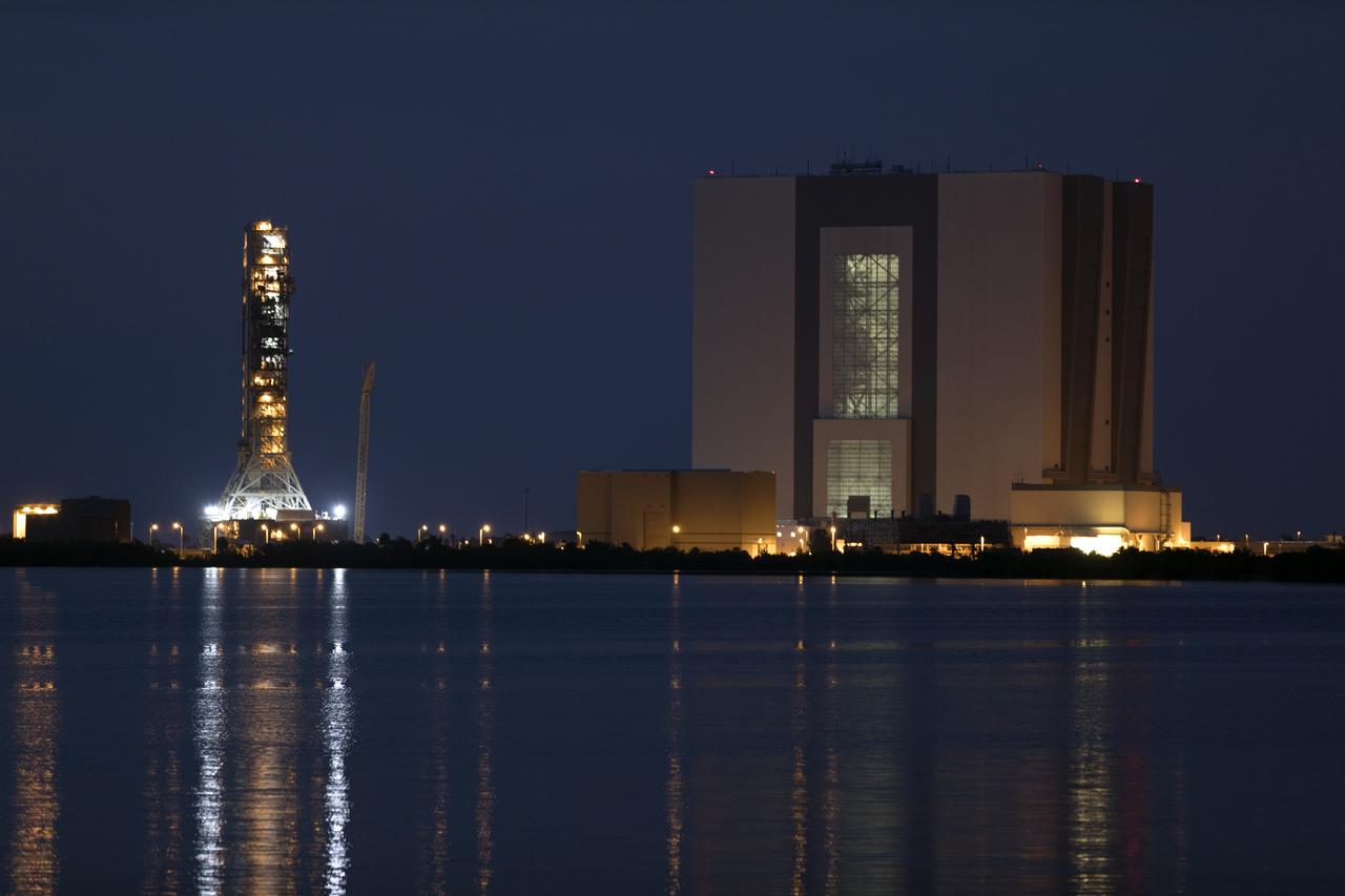 With their lights reflected in the water, a nighttime view of the iconic Vehicle Assembly Building, at right, and mobile launcher at NASA's Kennedy Space Center in Florida, on June 21, from the Saturn V center at nearby Kennedy Space Center Visitor Complex. Exploration Ground Systems is preparing both to support processing and launch of the agency's Space Launch System rocket and Orion spacecraft on Exploration Mission-1 and deep space missions.