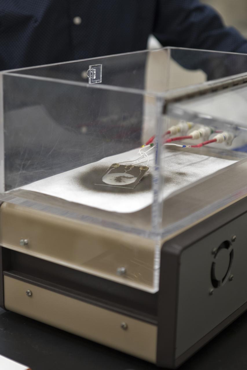 In the Electrostatics and Surface Physics Laboratory at NASA's Kennedy Space Center in Florida on Thursday, July 19, 2018, an experiment is underway in which an Electrostatic Dust Shield was been covered with dust similar to that which may be encountered by astronauts exploring the Moon or Mars. After activation, the device shakes off the dust. Scientists are developing the dust shield to help mitigate the problem of dust on equipment, astronauts' space suits and helmet visors. The device is slated for analysis aboard International Space Station t in the spring of 2019 o verify the effects of the space environment.