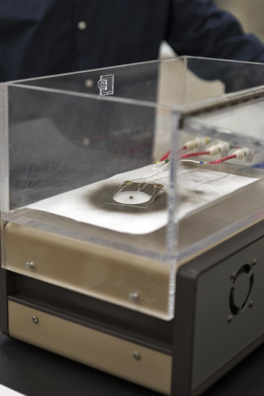 In the Electrostatics and Surface Physics Laboratory at NASA's Kennedy Space Center in Florida on Thursday, July 19, 2018, an experiment is underway in which an Electrostatic Dust Shield was been covered with dust similar to that which may be encountered by astronauts exploring the Moon or Mars. After activation, the device shakes off the dust. Scientists are developing the dust shield to help mitigate the problem of dust on equipment, astronauts' space suits and helmet visors. The device is slated for analysis aboard International Space Station in the spring of 2019 to verify the effects of the space environment.