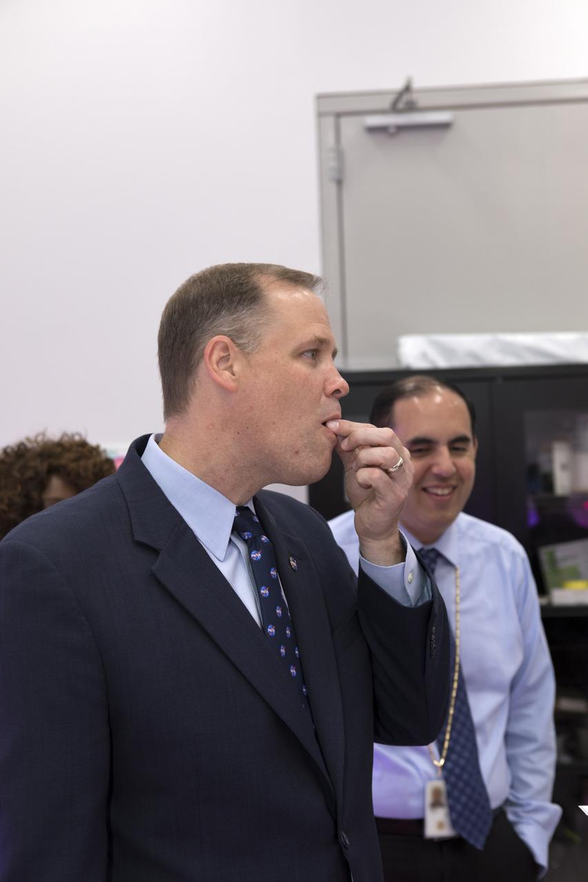 NASA Administrator Jim Bridenstine, left, tours a plant research laboratory and samples a microgreen inside the Space Station Processing Facility (SSPF) at NASA's Kennedy Space Center in Florida, on Aug. 7, 2018. Behind Bridenstine is Bryan Onate, Advanced Plant Habitat project manager. Bridenstine received updates on research and technology accomplishments during his visit to the SSPF.