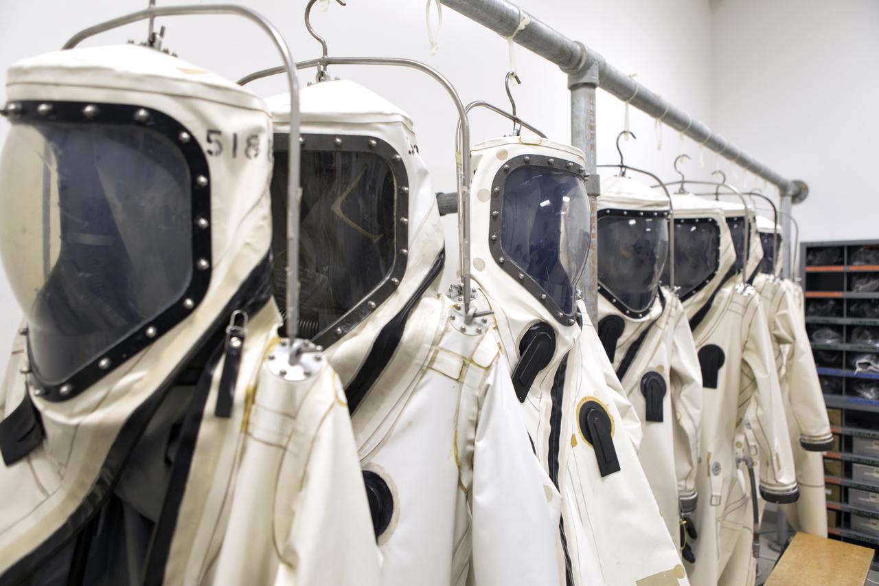 Self-Contained Atmospheric Protective Ensemble (SCAPE) suits are hanging in a row inside the Multi-Payload Processing Facility (MPPF) at NASA's Kennedy Space Center in Florida on Oct. 31, 2018. SCAPE operators will don the suits and then participate in a hypergolic systems hot flow test at the MPPF. The test will serve as operational validation of the hypergol subsystem and demonstrate that the hypergols subsystem can service the Orion spacecraft, flow fuel at the required rates, drain and de-service the system, and meet the intended timeline. SCAPE suite are used in operations involving toxic propellants and are supplied with air either through a hardline or through a self-contained environmental control unit.