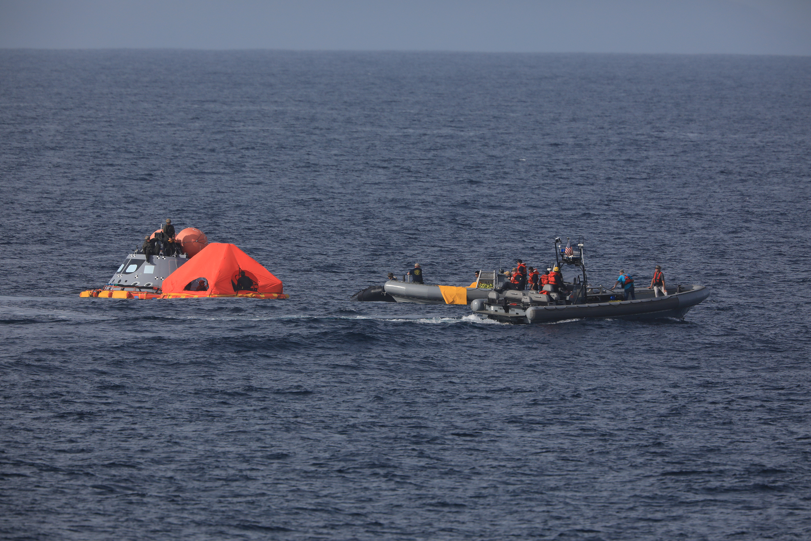 In the open ocean, a small boat converges on a mockup of the Orion capsule with a bright orange inflatable canopy in front of it.