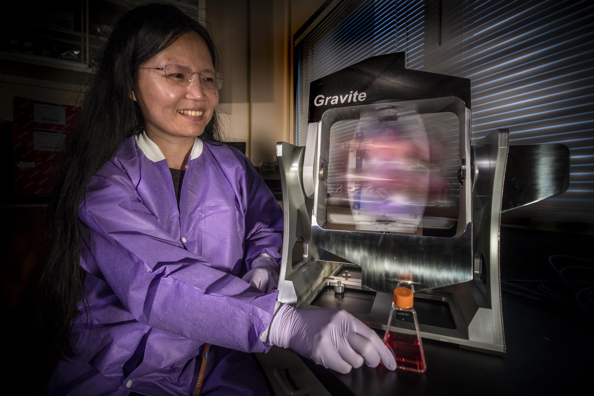 A female scientist in a purple lab coat smiles as the inserts a vial of liquid into a gray square-shaped microgravity machine.
