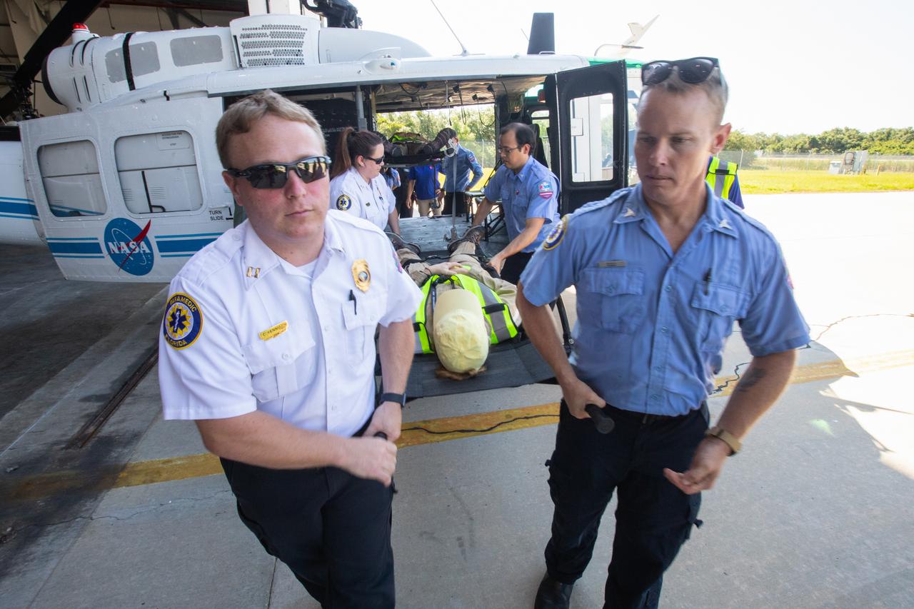 Kennedy Space Center personnel and American Medical Response (AMR) contractor paramedics gather around a “patient,” a KEMCON Fitness Center staff member, during a medical support training course in the Space Florida hangar at the spaceport’s Shuttle Landing Facility on May 17, 2019. The course was designed to familiarize the AMR paramedics with the center’s Triage Forces deployment, which included medical team members, fire/rescue personnel, environmental health specialists and flight operations crew members, as well as a helicopter, Mine Resistant Ambush Protected (MRAP) tactical vehicle, fire pumper truck and triage vehicles. The AMR paramedics will assist the agency in contingency planning for the return of human spaceflight from Kennedy.