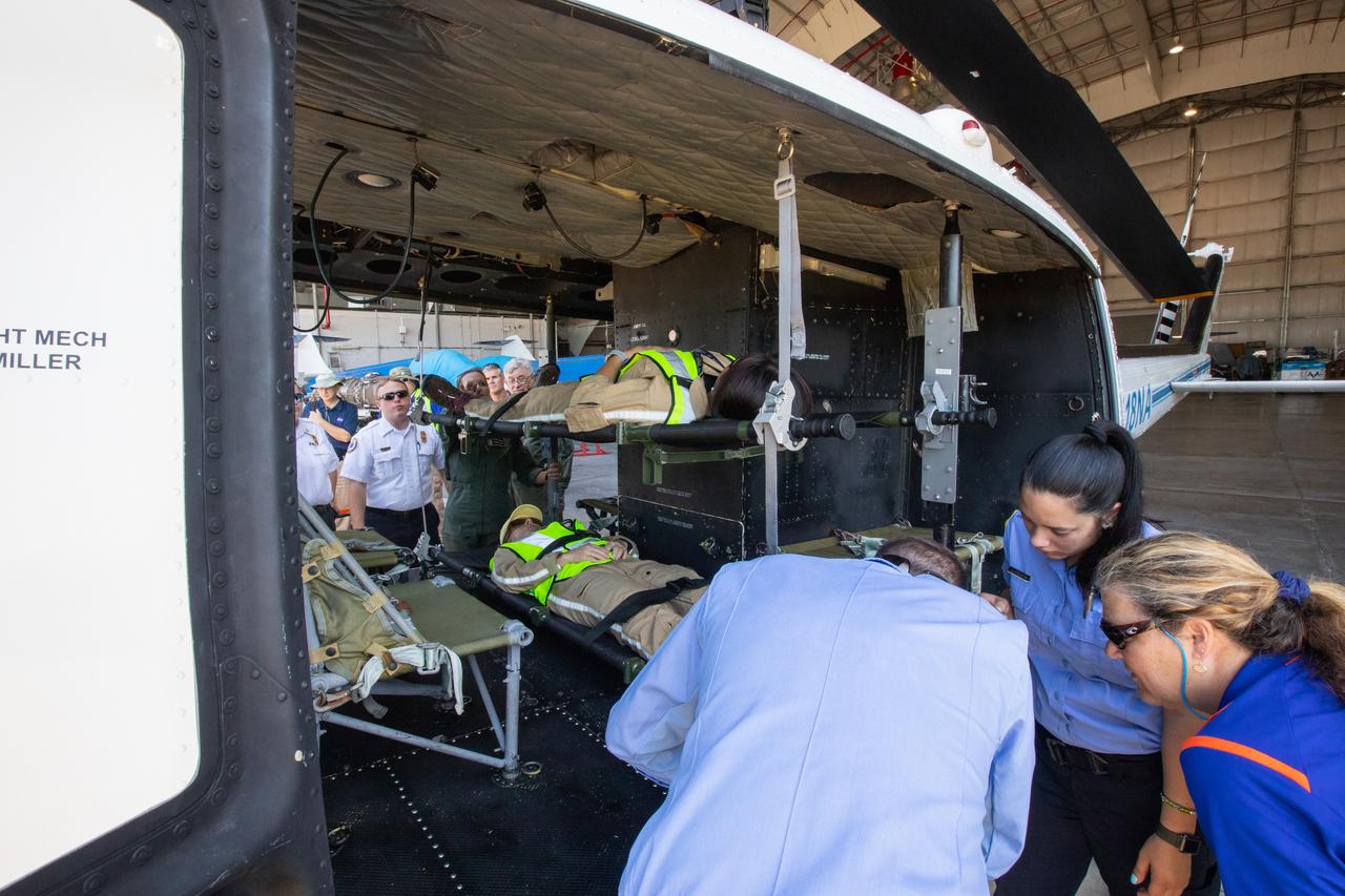 Kennedy Space Center personnel and American Medical Response (AMR) contractor paramedics gather around a “patient,” a KEMCON Fitness Center staff member, during a medical support training course in the Space Florida hangar at the spaceport’s Shuttle Landing Facility on May 17, 2019. The course was designed to familiarize the AMR paramedics with the center’s Triage Forces deployment, which included medical team members, fire/rescue personnel, environmental health specialists and flight operations crew members, as well as a helicopter, Mine Resistant Ambush Protected (MRAP) tactical vehicle, fire pumper truck and triage vehicles. The AMR paramedics will assist the agency in contingency planning for the return of human spaceflight from Kennedy.