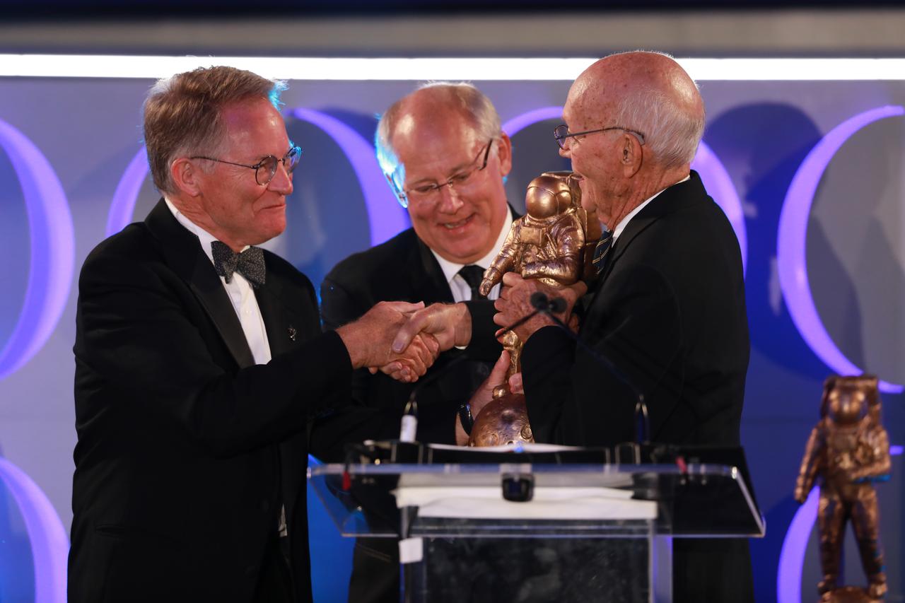 Michael Collins; right; Apollo 11 astronaut; presents the Innovation Award to David Thompson; left; founder; president and CEO of Orbital Sciences Corp (later known as Orbital ATK); during the Apollo 50th Gala on July 16; 2019. The event; presented by Northrop Grumman; was held inside the Apollo/Saturn V Center at the Kennedy Space Center Visitor Comples in Florida. In the center at the podium is Andy Aldrin; son of Apollo 11 astronaut Buzz Aldrin.