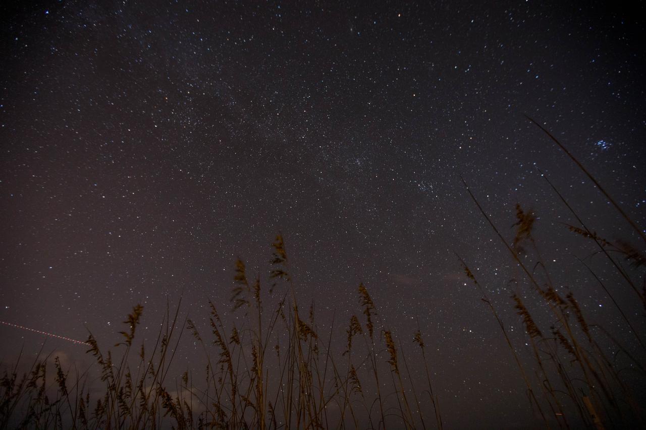 Sea oats frame the night sky, bisected by the Milky Way, in this view from the Atlantic shoreline at NASA’s Kennedy Space Center in Florida on Oct. 31, 2019. The lights of an aircraft create an illuminated streak in the lower left corner.