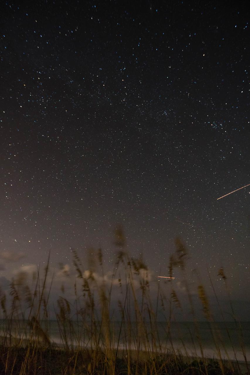Sea oats are backdropped by the night sky and the Milky Way in this view from the Atlantic shoreline at NASA’s Kennedy Space Center in Florida on Oct. 31, 2019. Lights from aircraft create illuminated streaks above the horizon and at right.