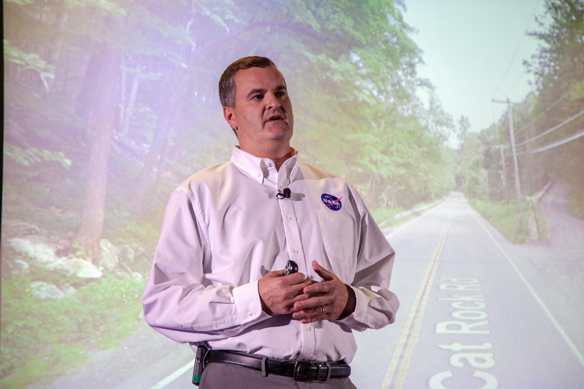 Speaker wearing a white shirt with a NASA logo, presenting in front of a projected image of a rural road surrounded by greenery. The speaker is gesturing with both hands while wearing a microphone headset, suggesting a professional talk or presentation.
