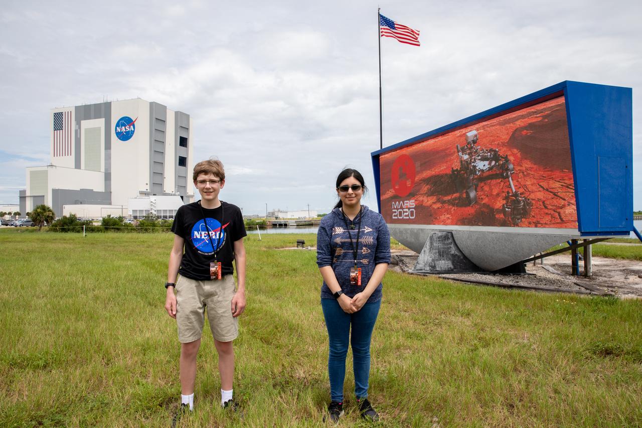 Students Alex Mather, at left, and Vaneeza Rupani, stand near the countdown clock at the News Center at NASA’s Kennedy Space Center in Florida on July 28, 2020. Mather named the Perseverance rover, and Rupani named the Ingenuity helicopter. In view in the background is the iconic Vehicle Assembly Building. The rover is scheduled to launch July 30, on a United Launch Alliance Atlas V 541 rocket from Space Launch Complex 41 at nearby Cape Canaveral Air Force Station. The rover is part of NASA’s Mars Exploration Program, a long-term effort of robotic exploration of the Red Planet. The rover will search for habitable conditions in the ancient past and signs of past microbial life on Mars. The Launch Services Program at Kennedy is responsible for launch management.