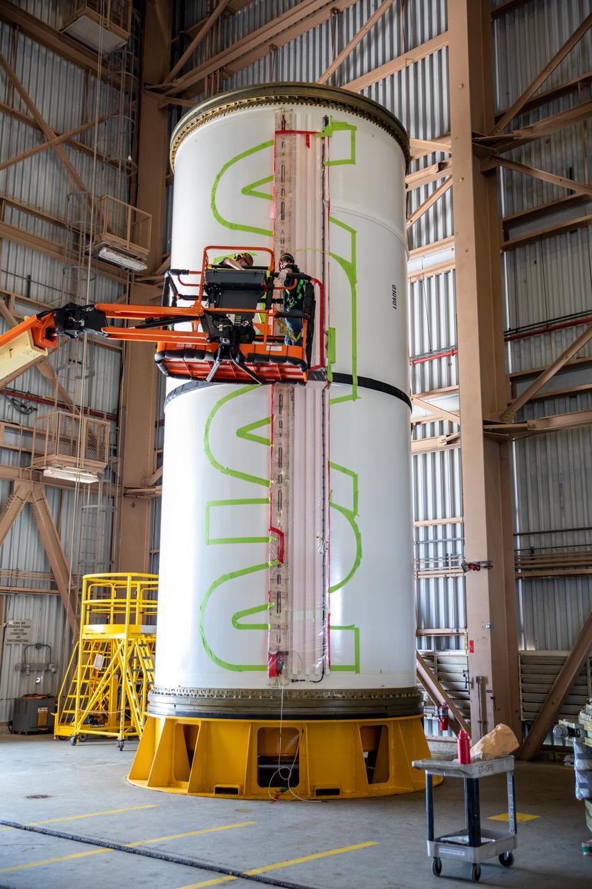 Workers with NASA’s Exploration Ground Systems (EGS) use a laser projector and green tape to mask off the shape of the agency’s “worm” logo on the side of an Artemis I solid rocket booster segment inside the Rotation, Processing and Surge Facility (RPSF) at Kennedy Space Center in Florida, Sept. 3, 2020. The booster segments will help propel the Space Launch System (SLS) rocket on Artemis I, a test of the Orion spacecraft and SLS as an integrated system ahead of crewed flights to the Moon. Northrop Grumman, which built the booster segments, is covering the cost of the painting.