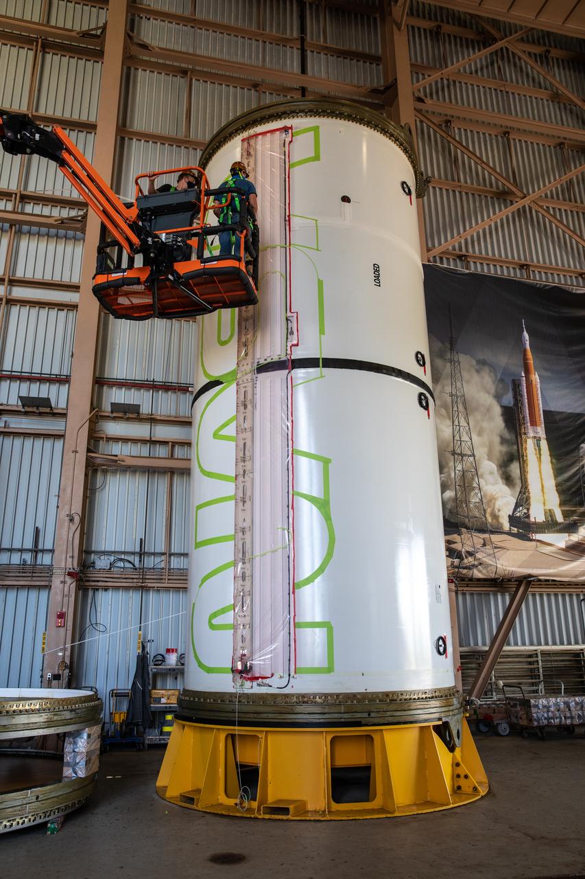 Workers with NASA’s Exploration Ground Systems (EGS) use a laser projector and green tape to mask off the shape of the agency’s “worm” logo on the side of an Artemis I solid rocket booster segment inside the Rotation, Processing and Surge Facility (RPSF) at Kennedy Space Center in Florida, Sept. 3, 2020. The booster segments will help propel the Space Launch System (SLS) rocket on Artemis I, a test of the Orion spacecraft and SLS as an integrated system ahead of crewed flights to the Moon. Northrop Grumman, which built the booster segments, is covering the cost of the painting.
