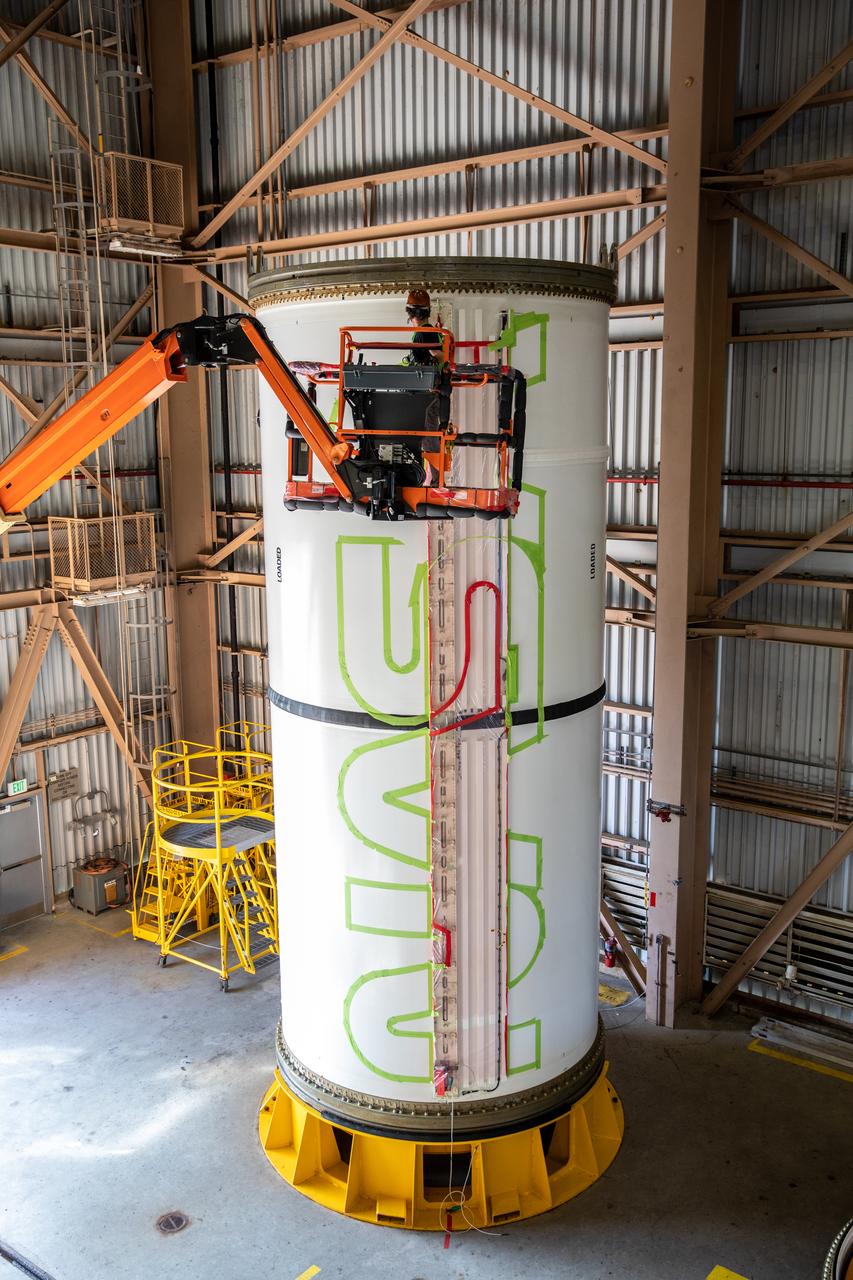 A worker with NASA’s Exploration Ground Systems (EGS) applies bright red paint to the agency’s “worm” logo taking shape on the side of an Artemis I solid rocket booster segment inside the Rotation, Processing and Surge Facility (RPSF) at Kennedy Space Center in Florida. The EGS team used a laser projector to mask off the logo with tape, then painted the first coat of the iconic design. The booster segments will help propel the Space Launch System (SLS) rocket on Artemis I, a test of the Orion spacecraft and SLS as an integrated system ahead of crewed flights to the Moon. Northrop Grumman, which built the booster segments, is covering the cost of the painting.