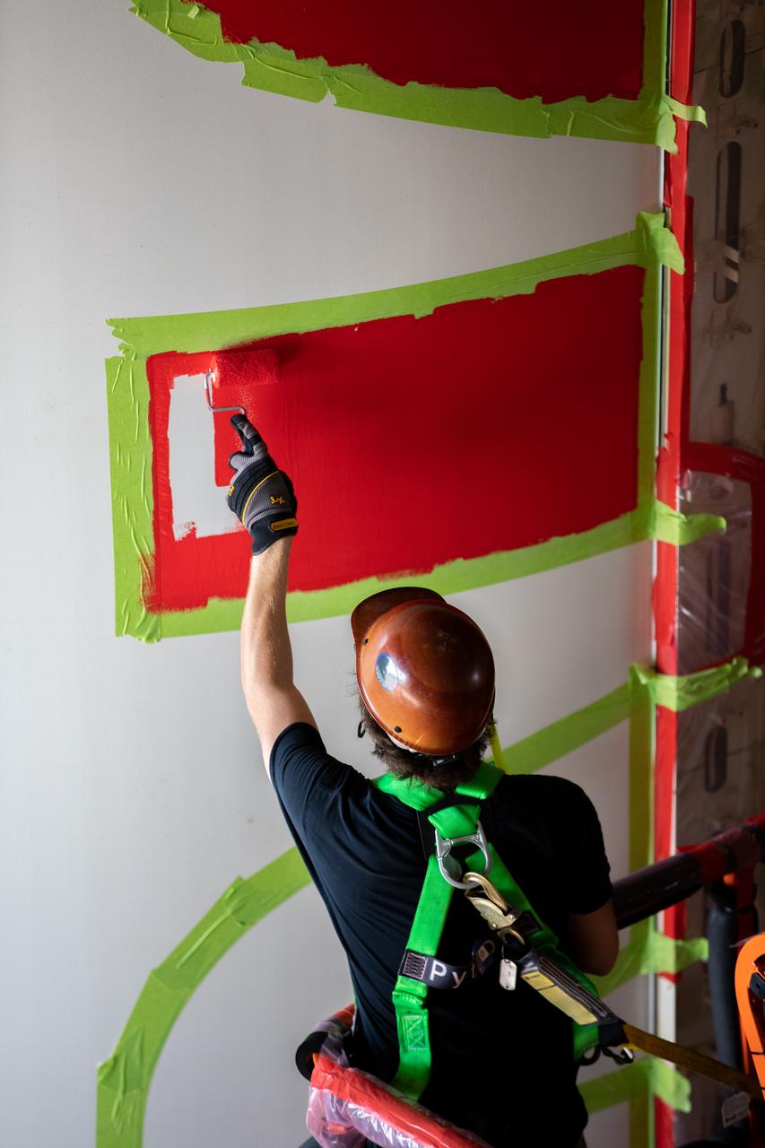 A worker with NASA’s Exploration Ground Systems (EGS) applies bright red paint to the agency’s “worm” logo taking shape on the side of an Artemis I solid rocket booster segment inside the Rotation, Processing and Surge Facility (RPSF) at Kennedy Space Center in Florida. The EGS team used a laser projector to mask off the logo with tape, then painted the first coat of the iconic design. The booster segments will help propel the Space Launch System (SLS) rocket on Artemis I, a test of the Orion spacecraft and SLS as an integrated system ahead of crewed flights to the Moon. Northrop Grumman, which built the booster segments, is covering the cost of the painting.