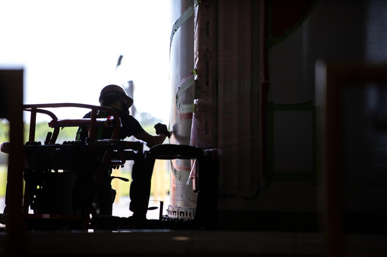 Silhouetted against the bright Florida sunlight outside, a worker with NASA’s Exploration Ground Systems (EGS) applies bright red paint to the agency’s “worm” logo taking shape on the side of an Artemis I solid rocket booster segment inside the Rotation, Processing and Surge Facility (RPSF) at Kennedy Space Center. The EGS team used a laser projector to mask off the logo with tape, then painted the first coat of the iconic design. The booster segments will help propel the Space Launch System (SLS) rocket on Artemis I, a test of the Orion spacecraft and SLS as an integrated system ahead of crewed flights to the Moon. Northrop Grumman, which built the booster segments, is covering the cost of the painting.