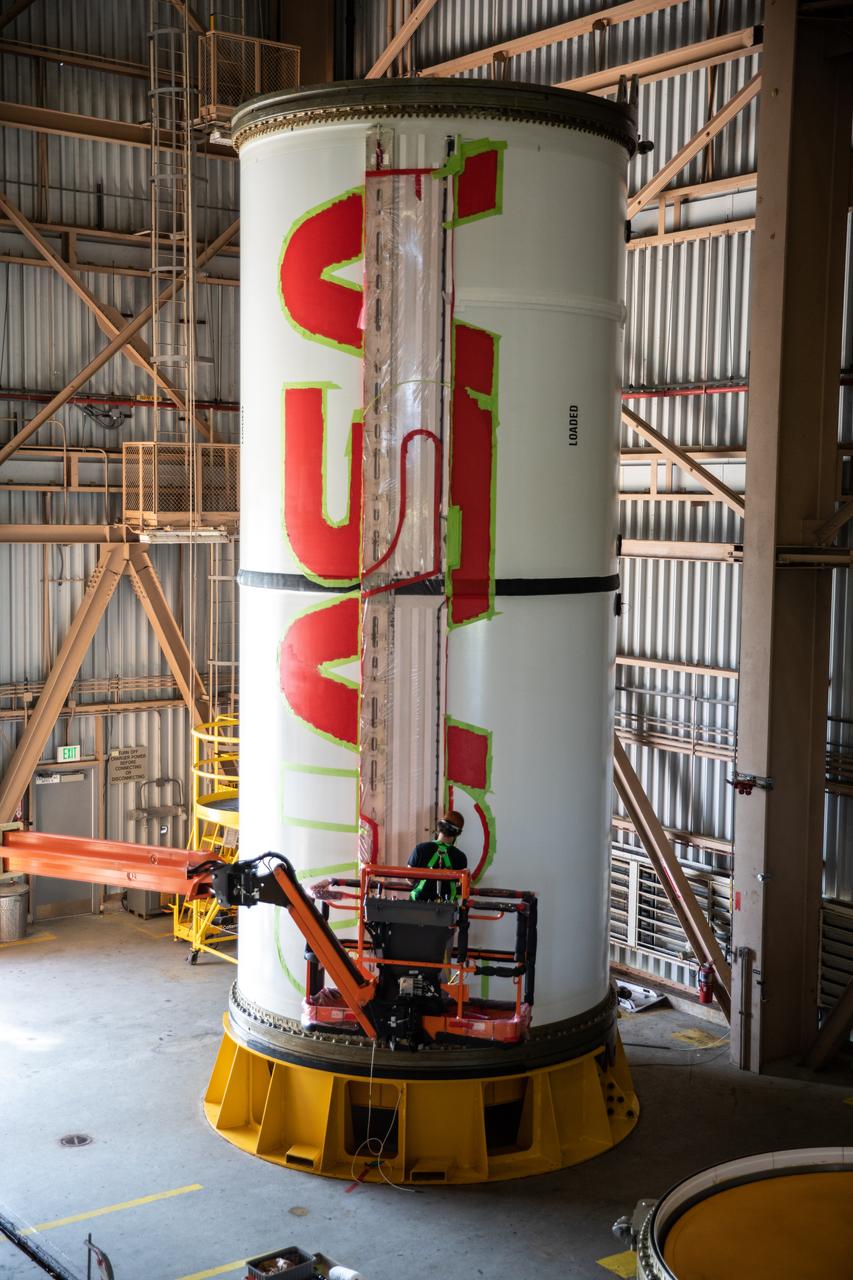 A worker with NASA’s Exploration Ground Systems (EGS) finishes the first coat of the bright red “worm” logo taking shape on the side of an Artemis I solid rocket booster segment inside the Rotation, Processing and Surge Facility (RPSF) at Kennedy Space Center in Florida. The EGS team used a laser projector to mask off the logo with tape, then painted the first coat of the iconic design. The booster segments will help propel the Space Launch System (SLS) rocket on Artemis I, a test of the Orion spacecraft and SLS as an integrated system ahead of crewed flights to the Moon. Northrop Grumman, which built the booster segments, is covering the cost of the painting.