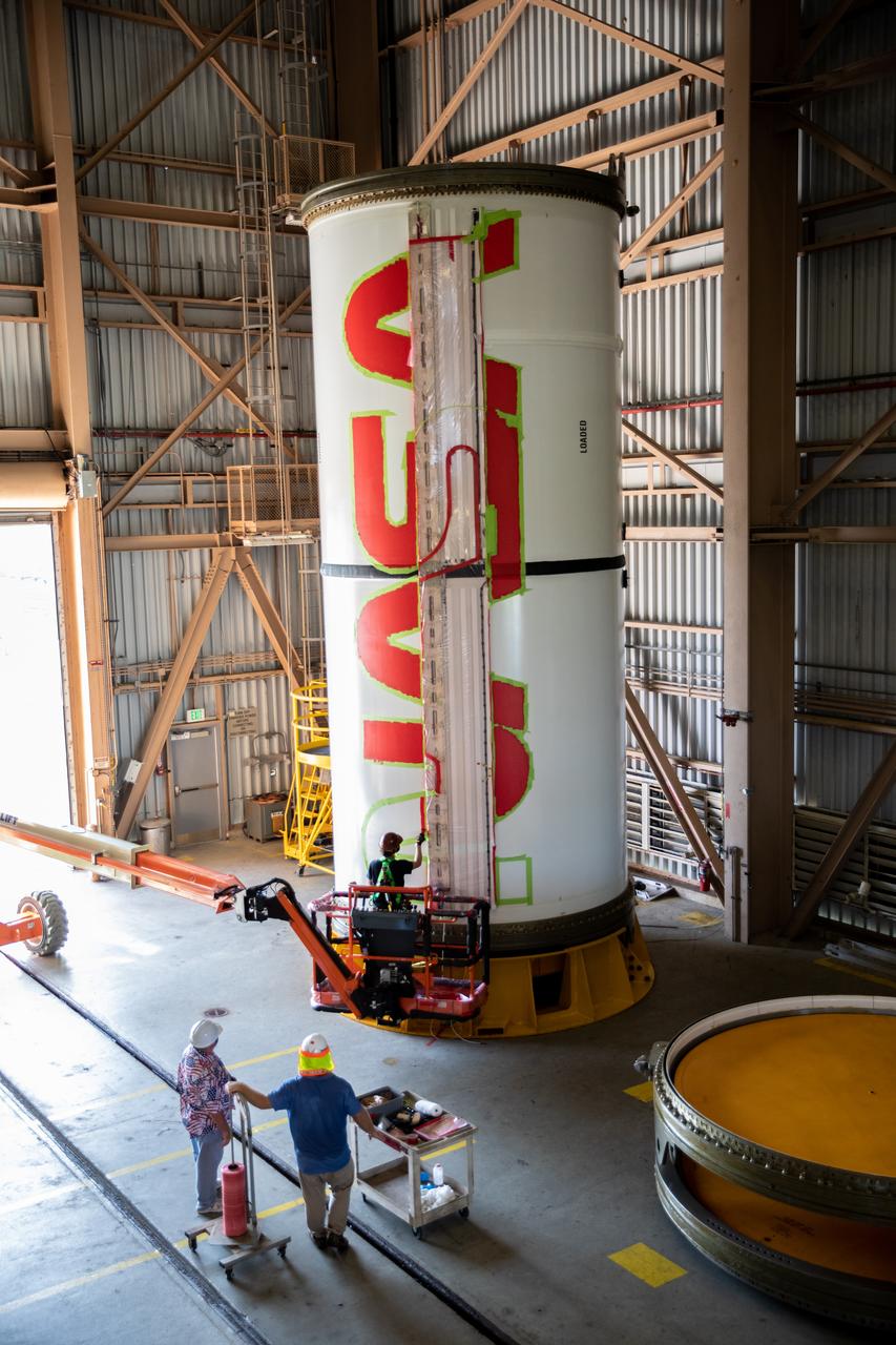 Workers with NASA’s Exploration Ground Systems (EGS) finishes the first coat of the bright red “worm” logo taking shape on the side of an Artemis I solid rocket booster segment inside the Rotation, Processing and Surge Facility (RPSF) at Kennedy Space Center in Florida. The EGS team used a laser projector to mask off the logo with tape, then painted the first coat of the iconic design. The booster segments will help propel the Space Launch System (SLS) rocket on Artemis I, a test of the Orion spacecraft and SLS as an integrated system ahead of crewed flights to the Moon. Northrop Grumman, which built the booster segments, is covering the cost of the painting.